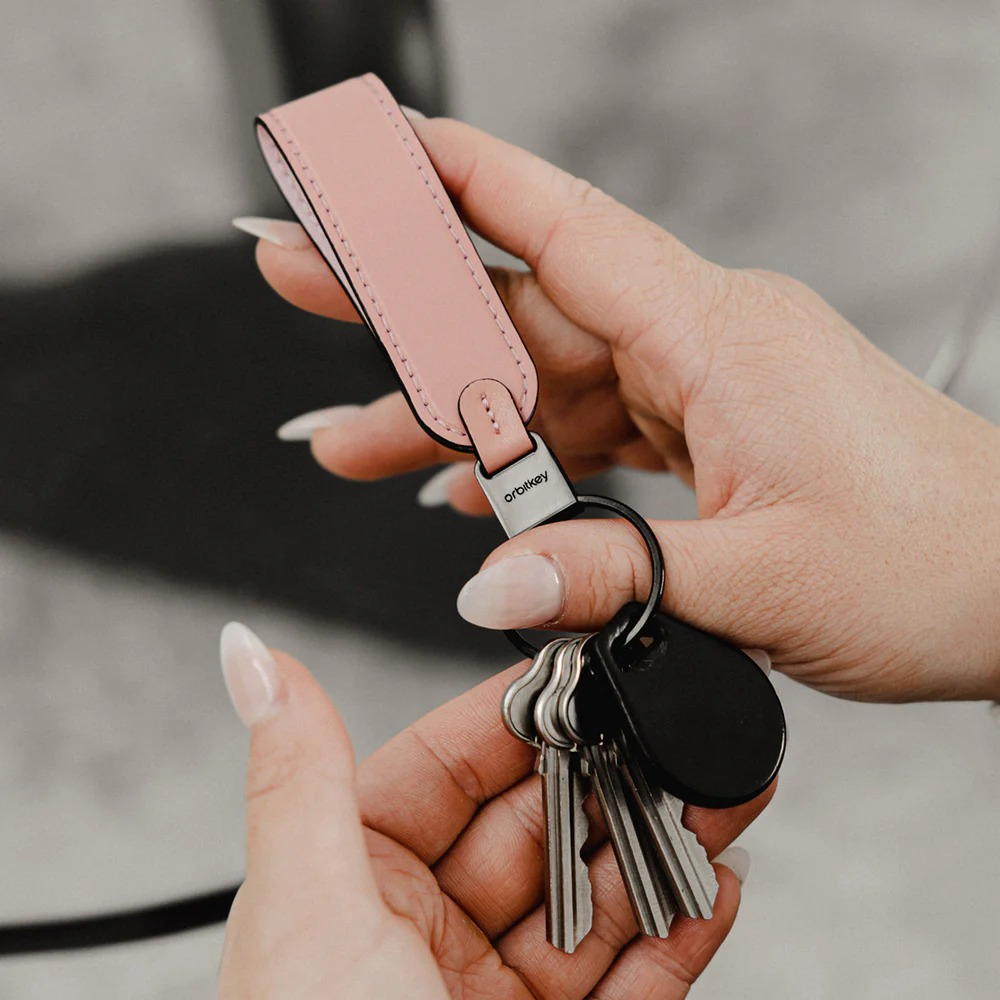 Ręka trzyma różowy skórzany pasek Orbitkey z metalowymi kluczami i czarnym brelokiem.