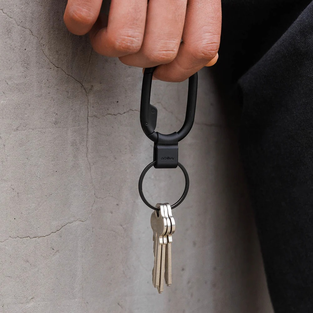 Ręka trzyma czarny karabińczyk i kółko Orbitkey z trzema srebrnymi kluczami na tle betonowej ściany.