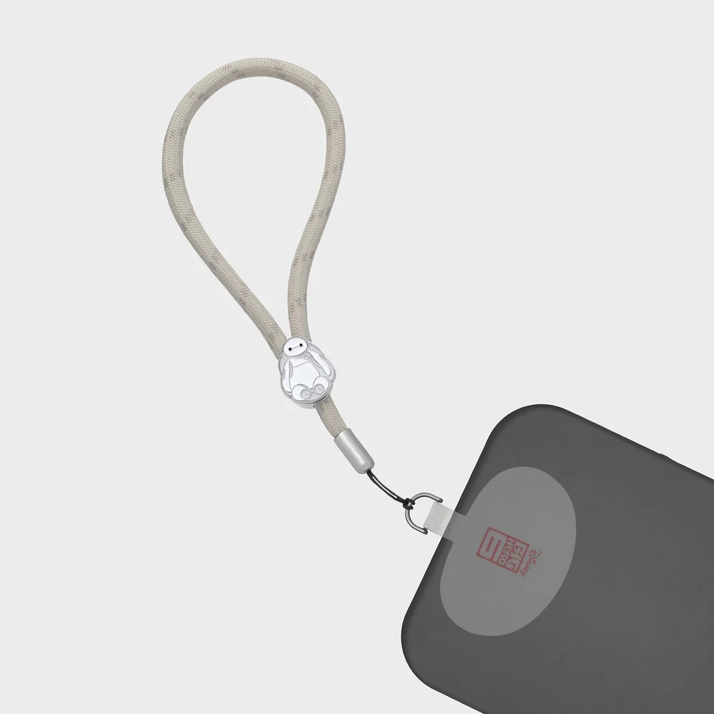 Beżowy pasek do telefonu z zawieszką Baymax przymocowany do ciemnego etui na jasnym tle.