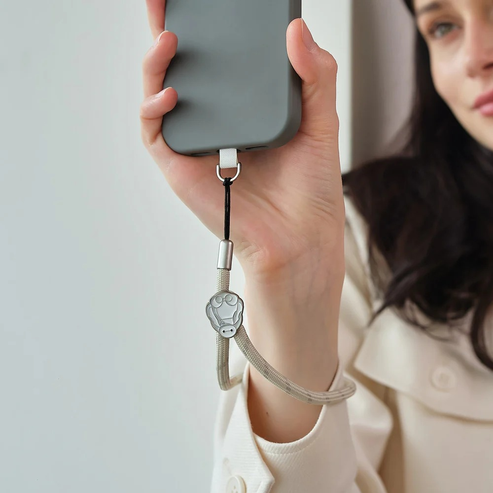 Kobieta trzyma szary smartfon z beżowym paskiem przymocowanym metalowym kółkiem i ozdobą z kreskówki.