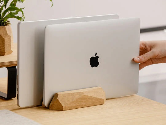 Dwa srebrne laptopy stoją pionowo w fasetowanym drewnianym stojaku na biurku.