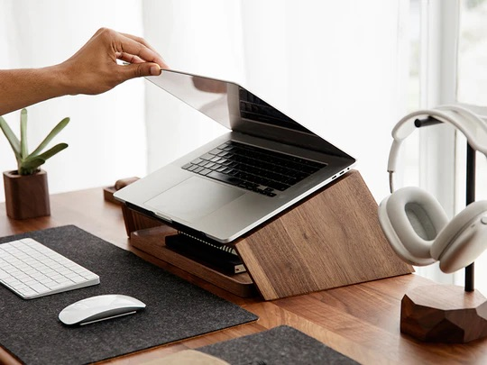 Ręka otwiera laptopa na drewnianym stojaku na biurku ze słuchawkami i klawiaturą.