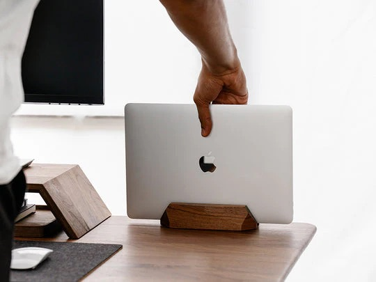 Ręka umieszcza srebrny laptop w drewnianym stojaku na biurku.