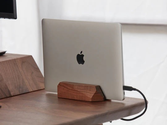 Zamknięty MacBook w drewnianym stojaku na drewnianym biurku, podłączony czarnym kablem.