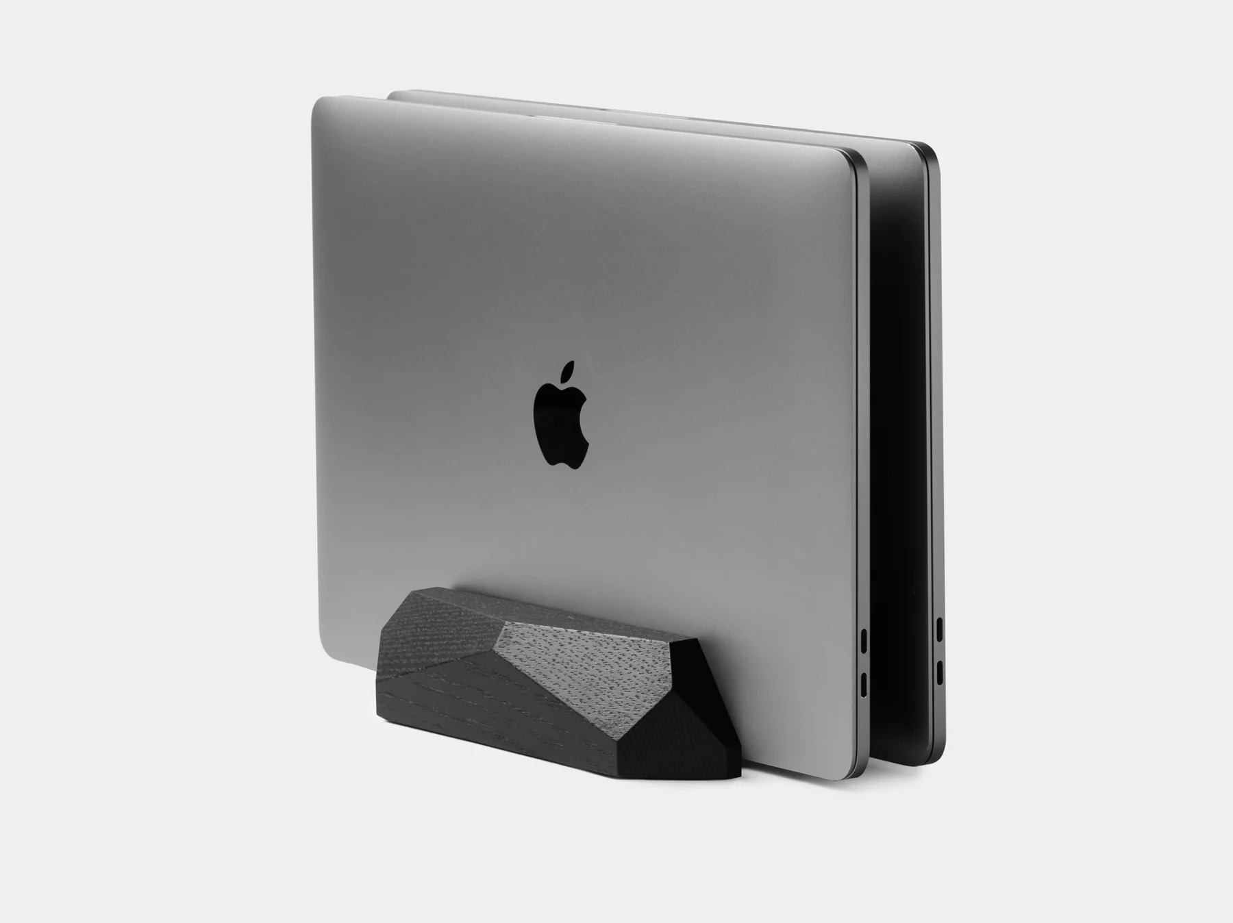 Dwa zamknięte szare MacBooki stoją pionowo w czarnym drewnianym stojaku na jasnym tle.