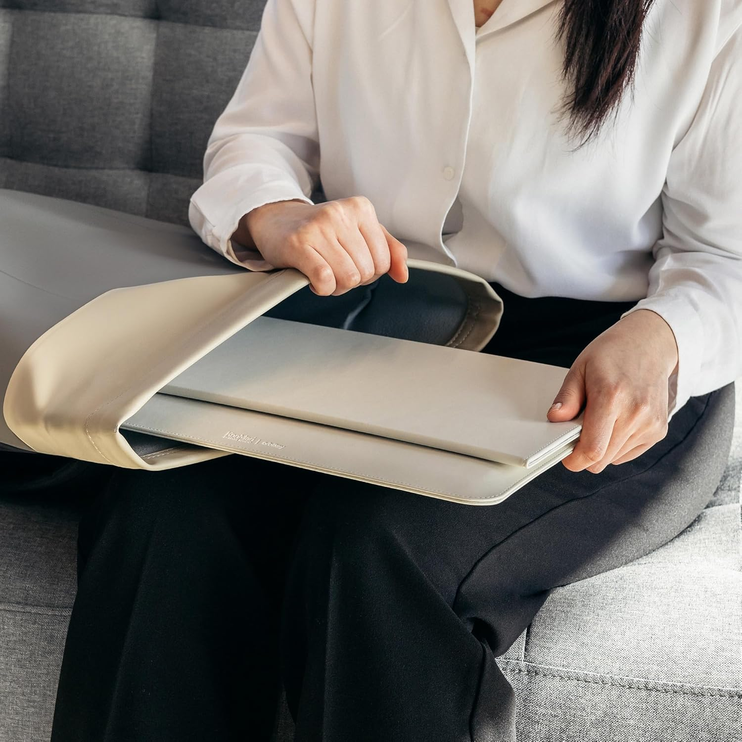 Kobieta w białej koszuli wkłada laptopa do kremowego etui na kolanach.