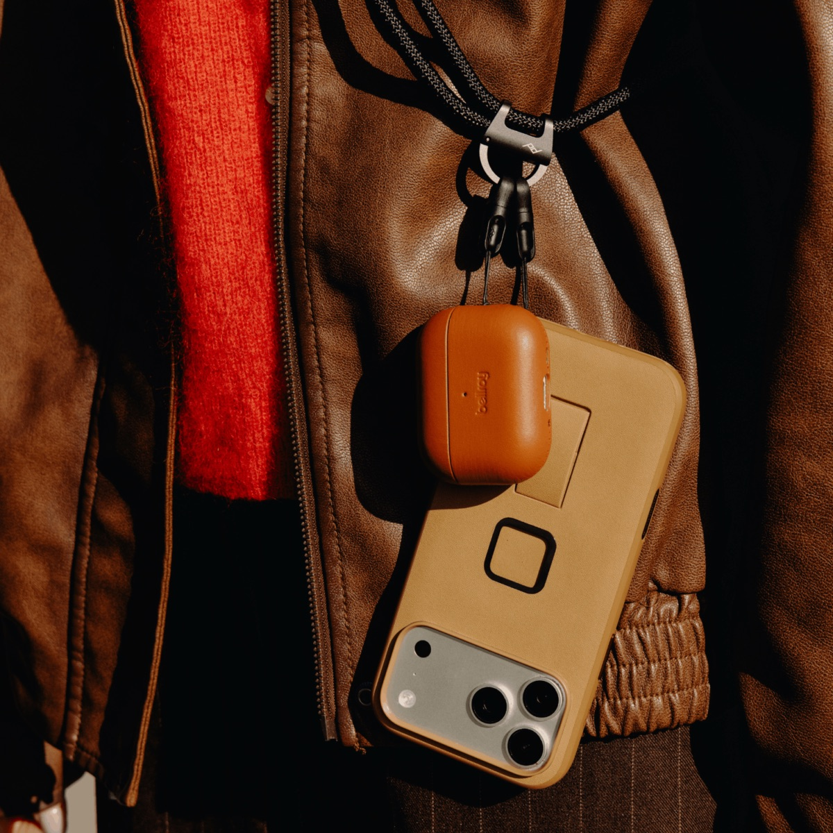 Brunatna kurtka skórzana, czerwony sweter, etui na telefon i AirPods wiszące na smyczy.
