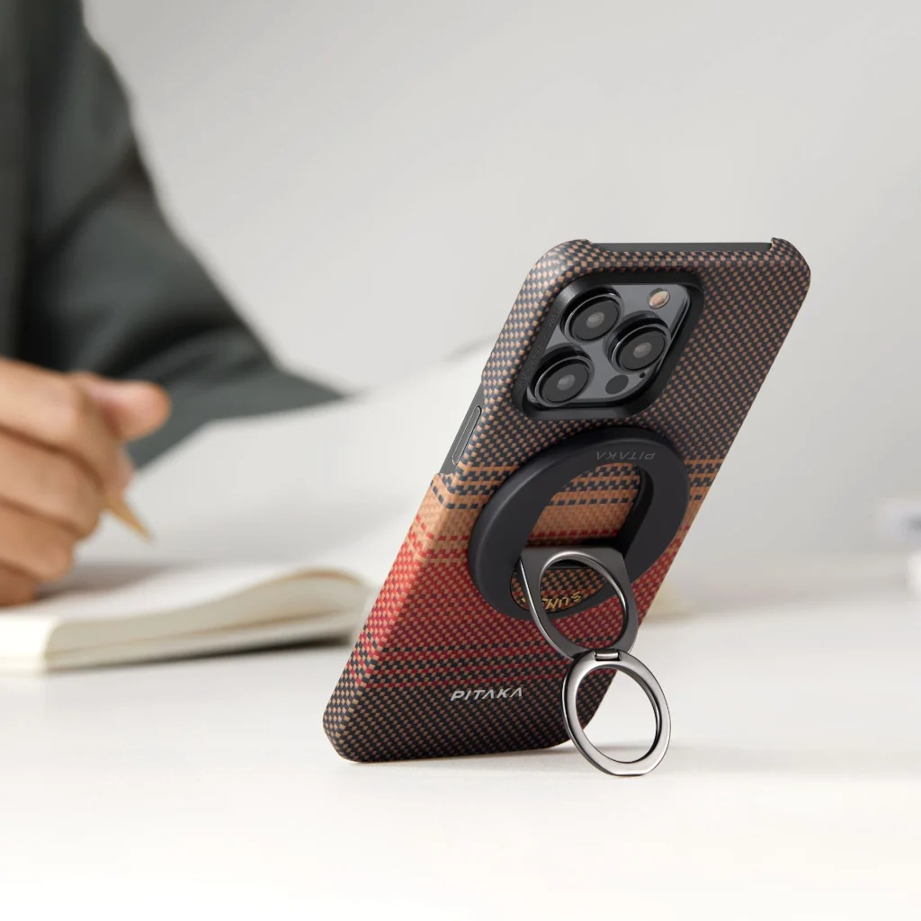 Wzorzyste etui na telefon z metalowym kółkiem stoi na białym biurku, gdy ktoś pisze w notesie.