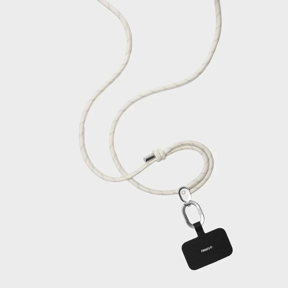Jasna smycz ze srebrnym zapięciem trzymająca małe czarne etui na telefon na szarym tle.