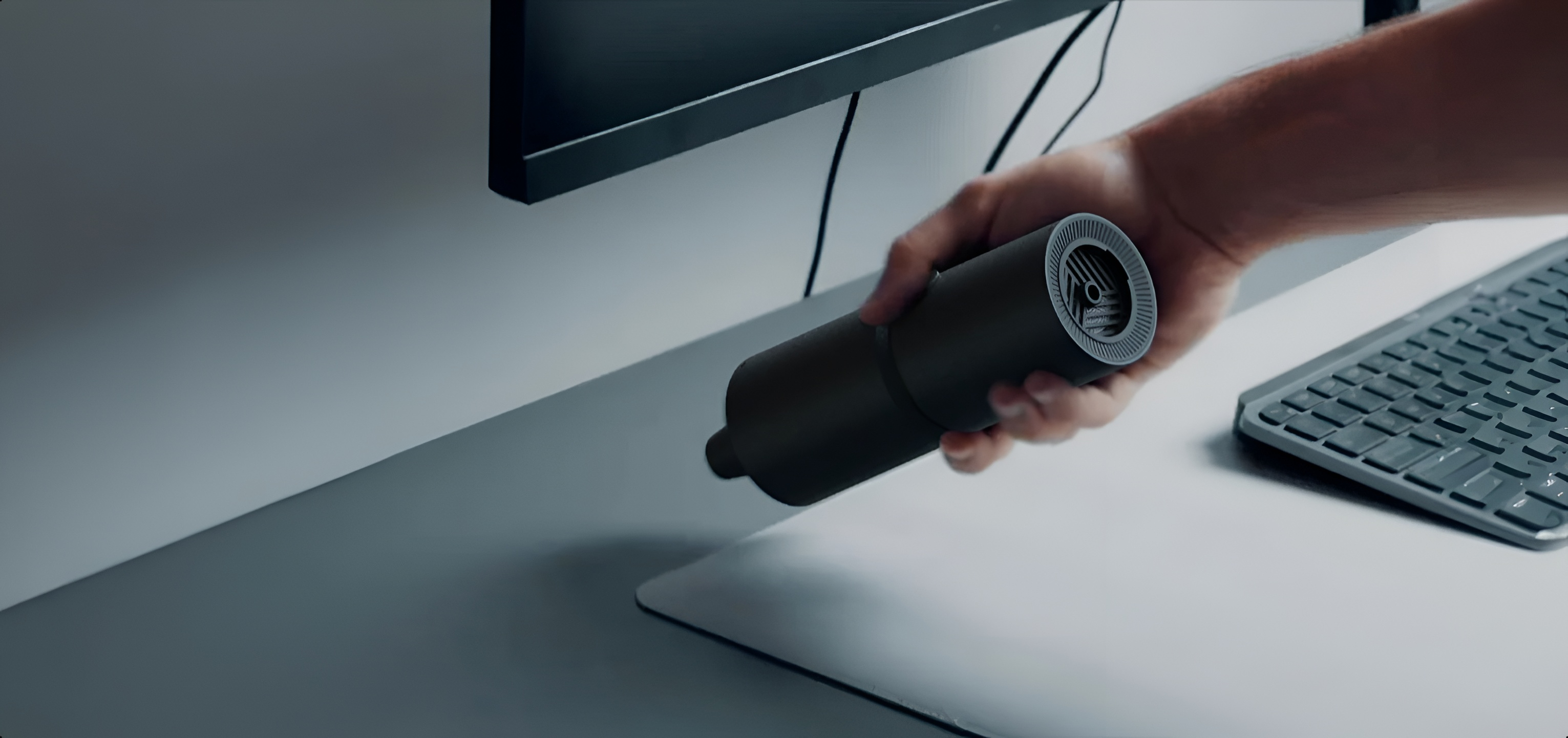 Ręka trzyma mały czarny cylinder nad biurkiem z monitorem i klawiaturą.