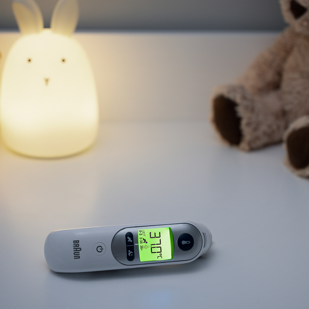 Cyfrowy termometr Braun leży na białej powierzchni, wskazując 37,0°C. Z tyłu świecąca lampka w kształcie królika i miś.