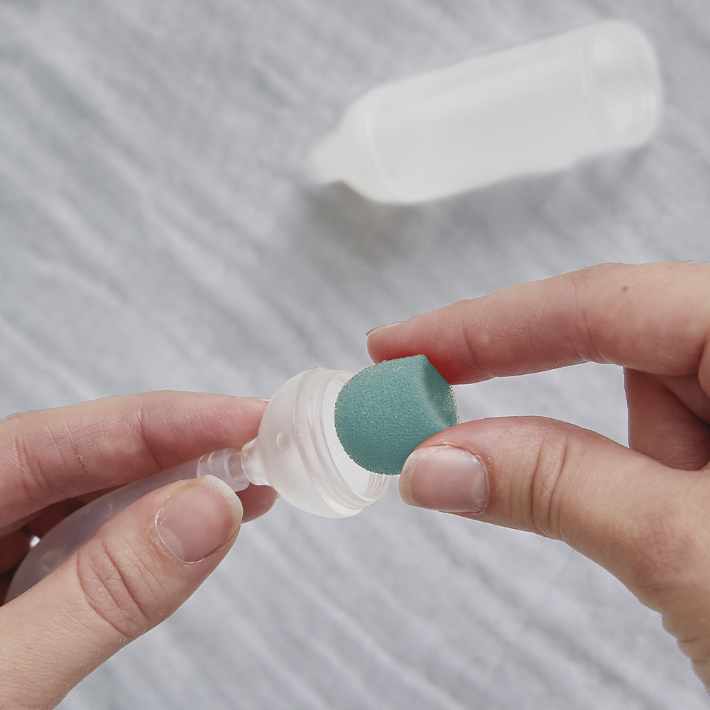 Ręce wkładają małą niebieską gąbkę do plastikowej rurki. W tle na białej tkaninie leży butelka.