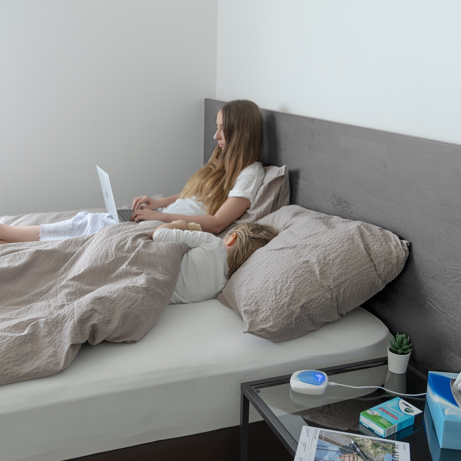 Kobieta pracuje na laptopie w łóżku, podczas gdy dziecko śpi obok niej.