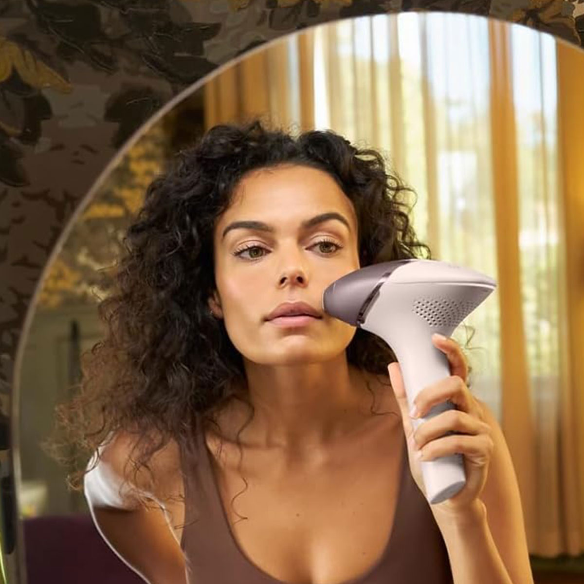 Donna con capelli ricci scuri tiene un dispositivo IPL bianco sul viso davanti a uno specchio.
