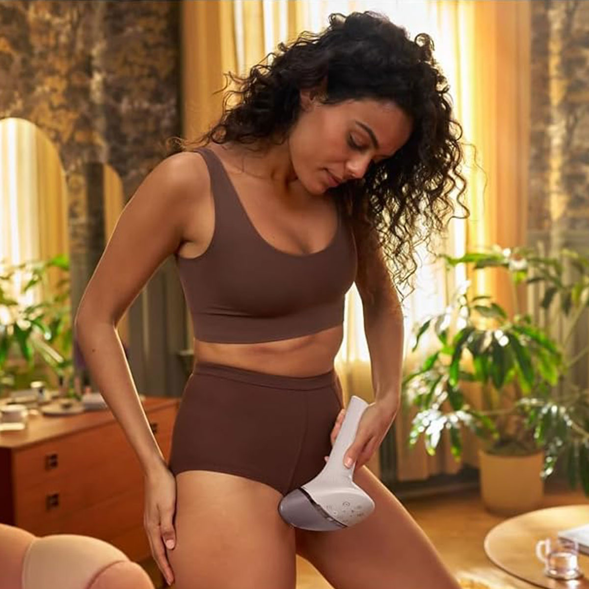 Una donna con capelli ricci scuri usa un depilatore sulla coscia, in interno con luce diurna.