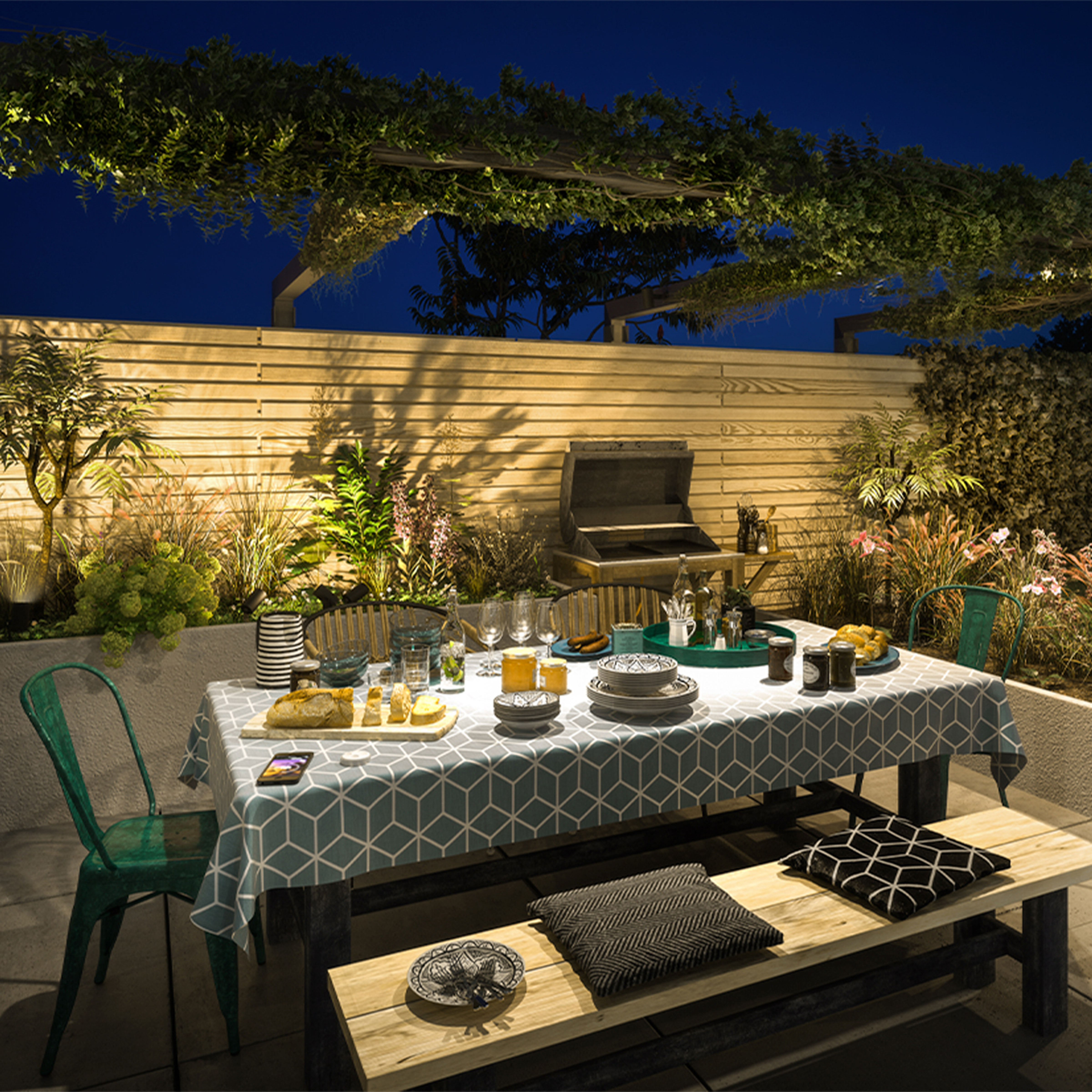 Beleuchtete Gartenterrasse nachts mit gedecktem Tisch, Holzbank, Grill und üppiger Bepflanzung.