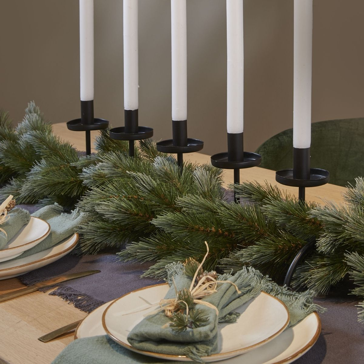 Table de fête avec des bougies blanches dans des supports noirs, guirlande de pin, assiettes et serviettes vertes.