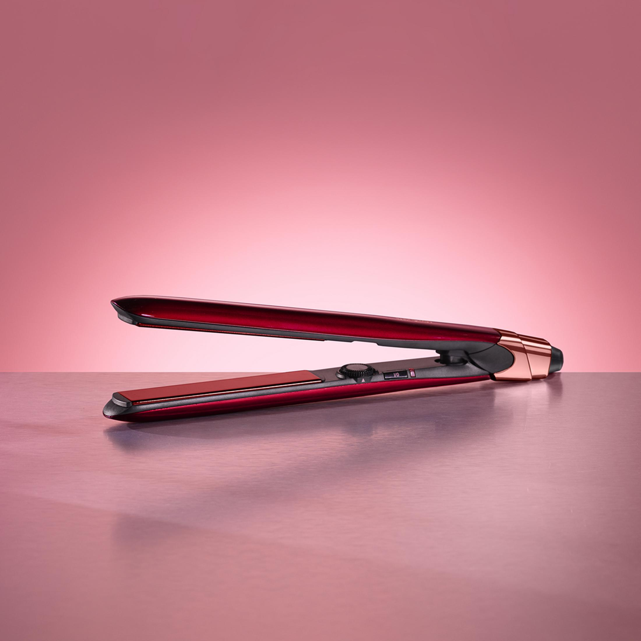 Una piastra per capelli rossa metallizzata poggia su una superficie rosa riflettente con sfondo rosa.