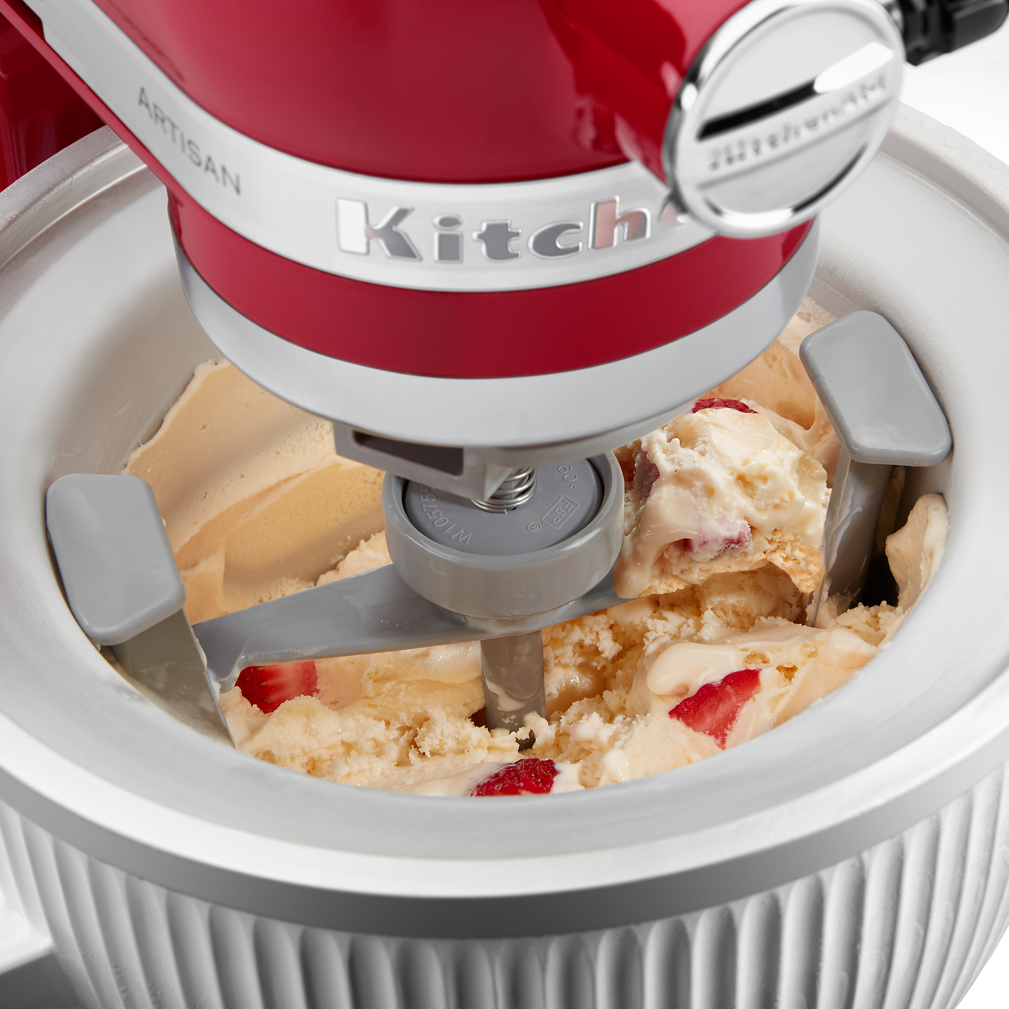 Gelatiera rossa KitchenAid con pala grigia che mescola gelato cremoso con pezzi di fragola in una ciotola bianca.