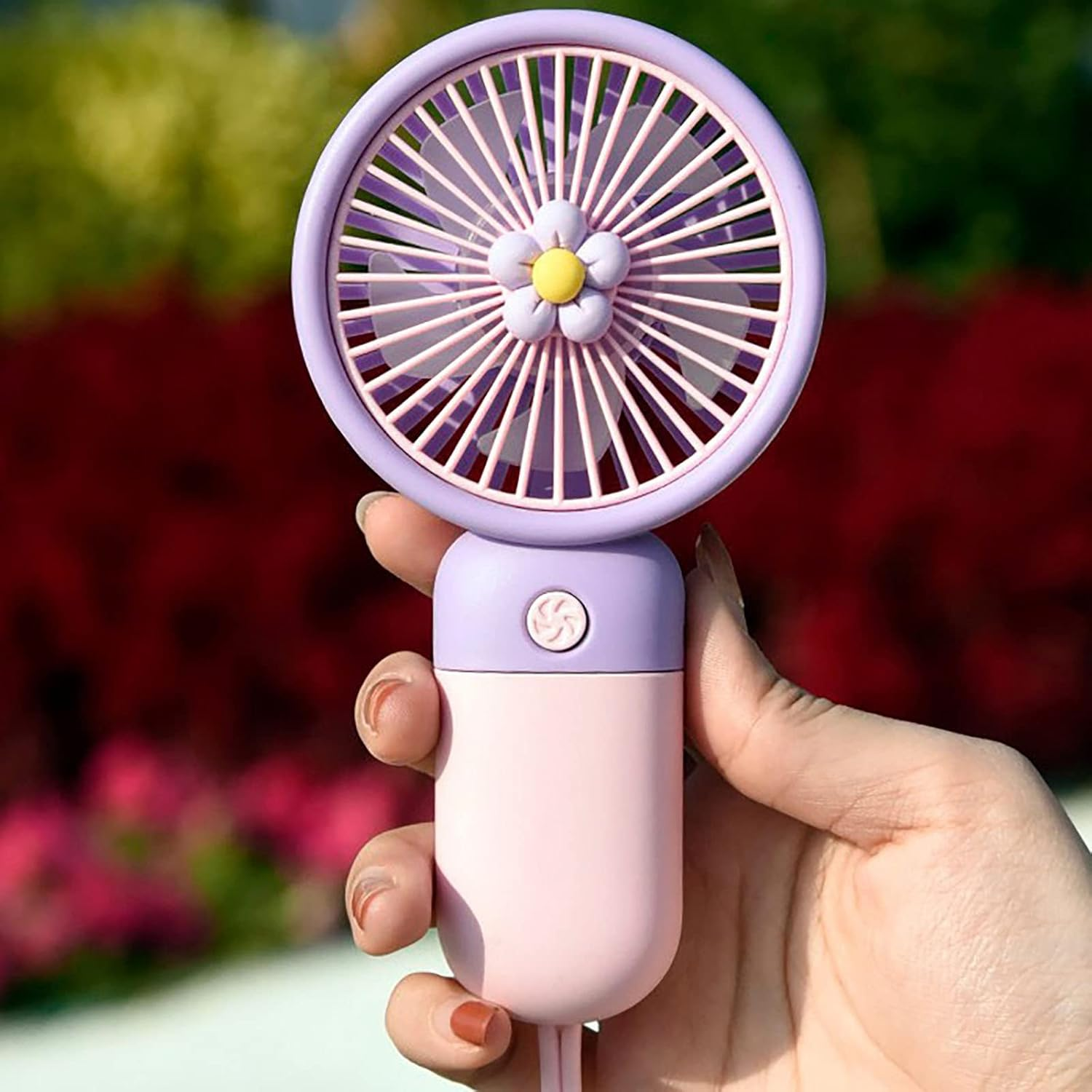 Eine Hand hält einen kleinen, runden, lilafarbenen und rosafarbenen Handventilator vor grünen und roten Blumen.