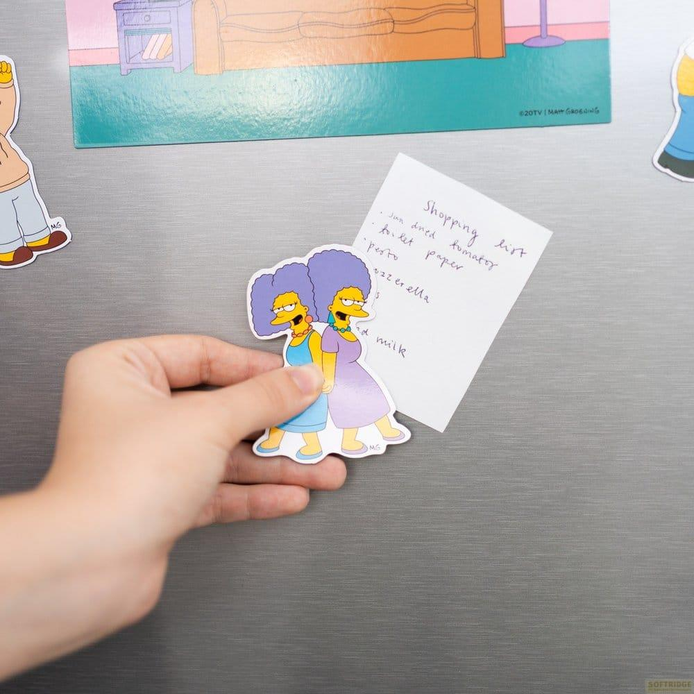 Hand hält Marge-Simpson-Magneten neben Einkaufsliste auf Kühlschrank.