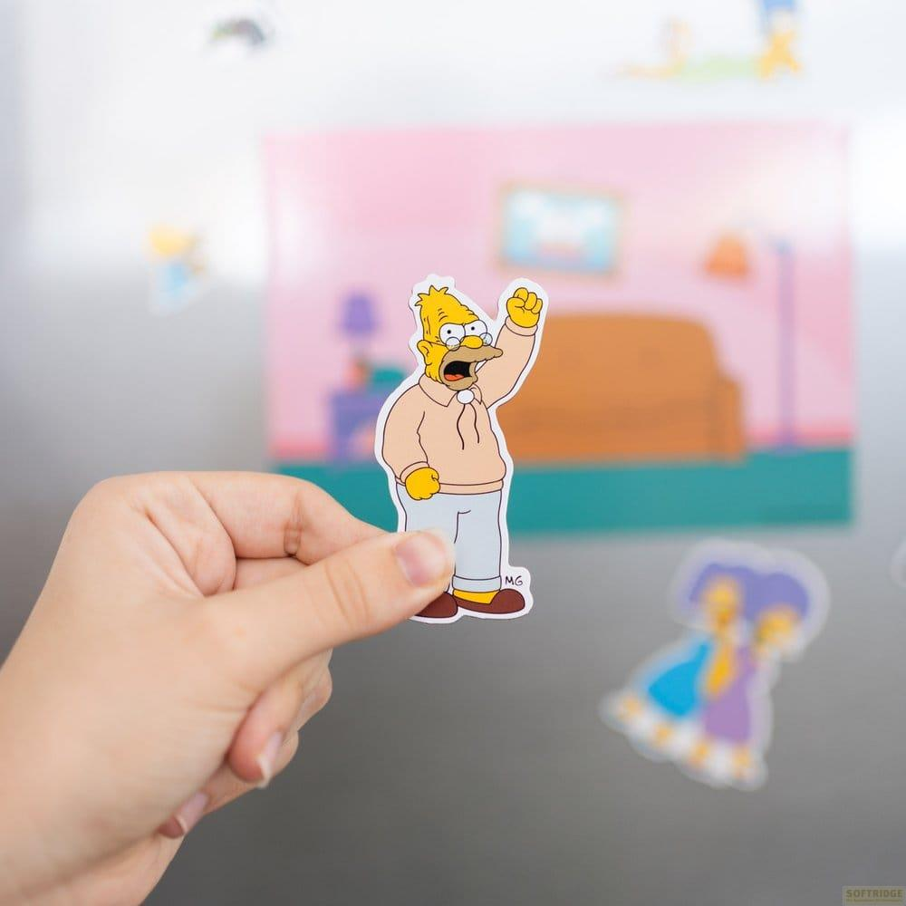 Eine Hand hält einen Sticker von Homer Simpson mit erhobener Faust vor einem Wohnzimmerhintergrund.
