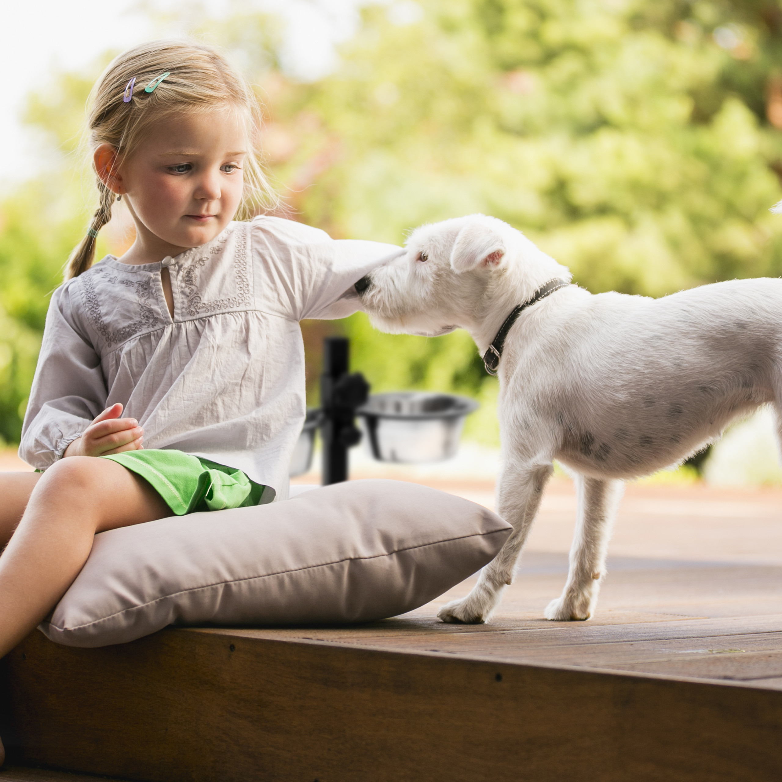 Ein blondes Mädchen im weißen Hemd sitzt auf einer Decke und streichelt einen kleinen weißen Hund auf einer Holzterrasse.