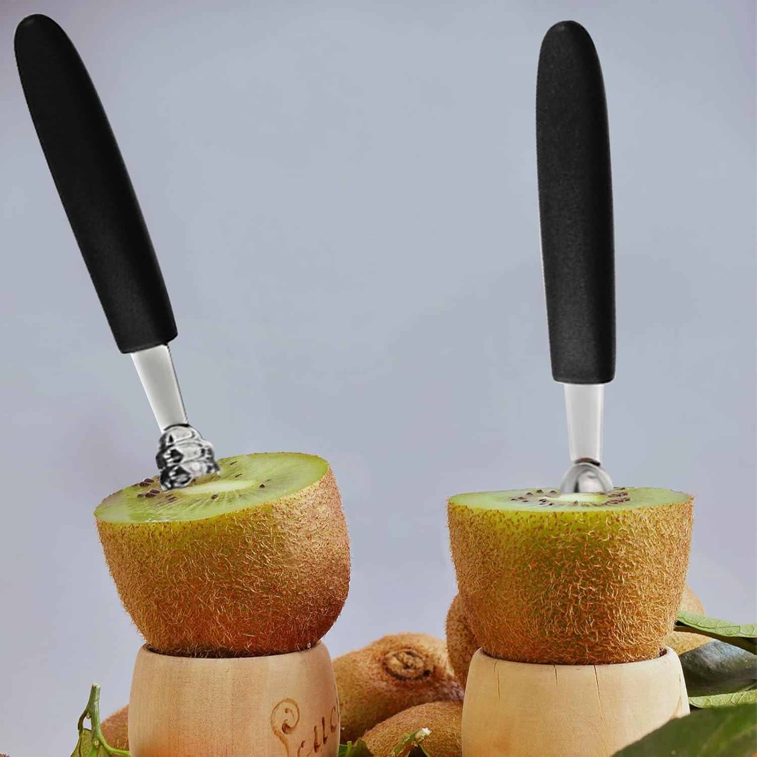 Zwei Kiwi-Hälften in Holzbechern mit Werkzeugen zum Herausschneiden des Fruchtfleischs vor grauem Hintergrund.