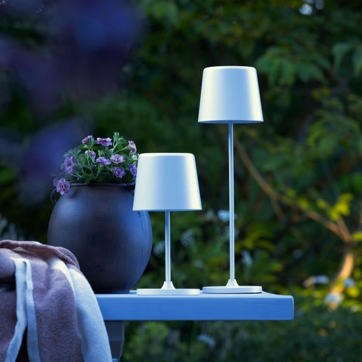 Zwei weiße Tischlampen, ein Pflanztopf mit violetten Blüten und eine Decke stehen auf einem Tisch im Freien.