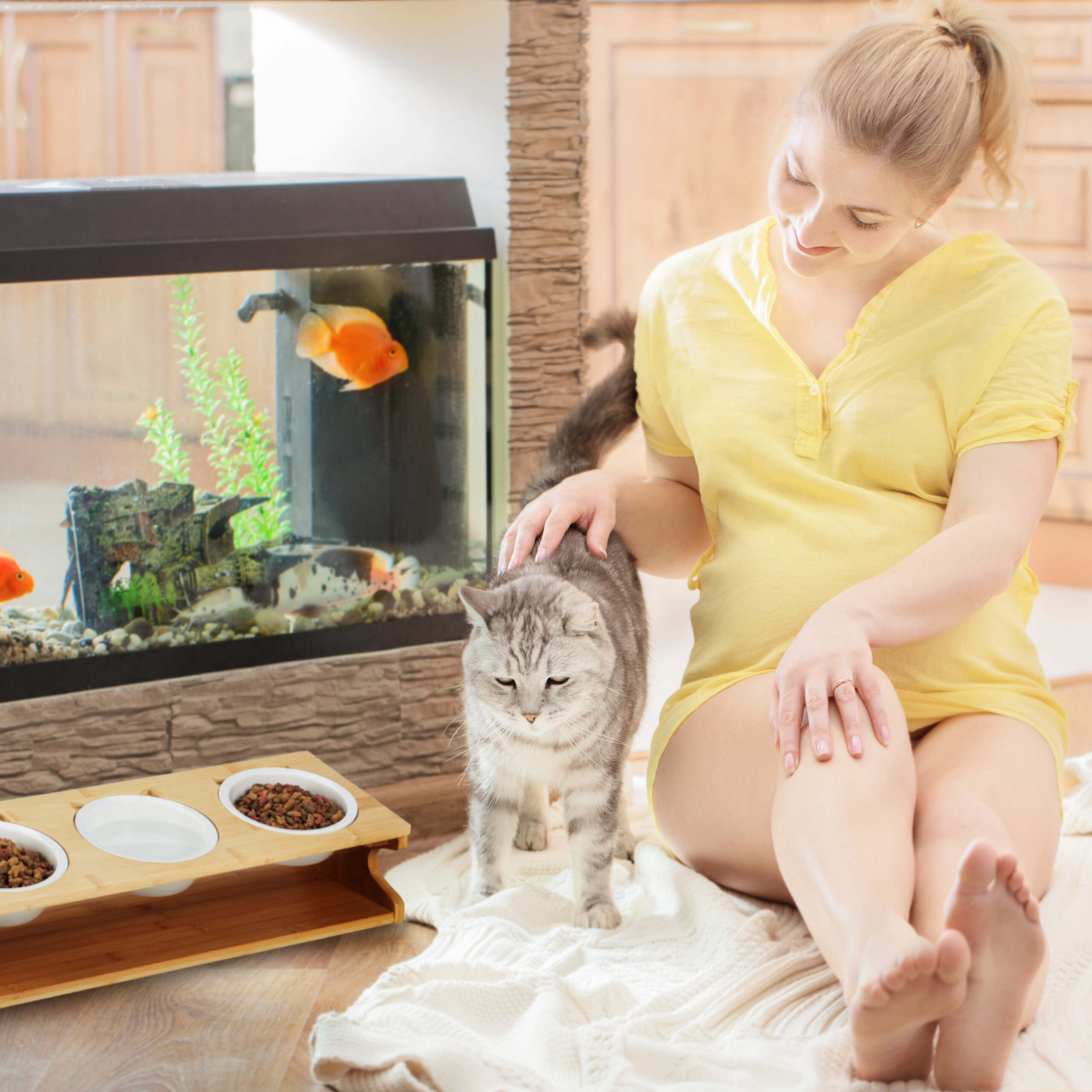 Eine blonde Frau im gelben Oberteil sitzt auf einem Teppich, streichelt eine graue Katze, links steht ein Aquarium.