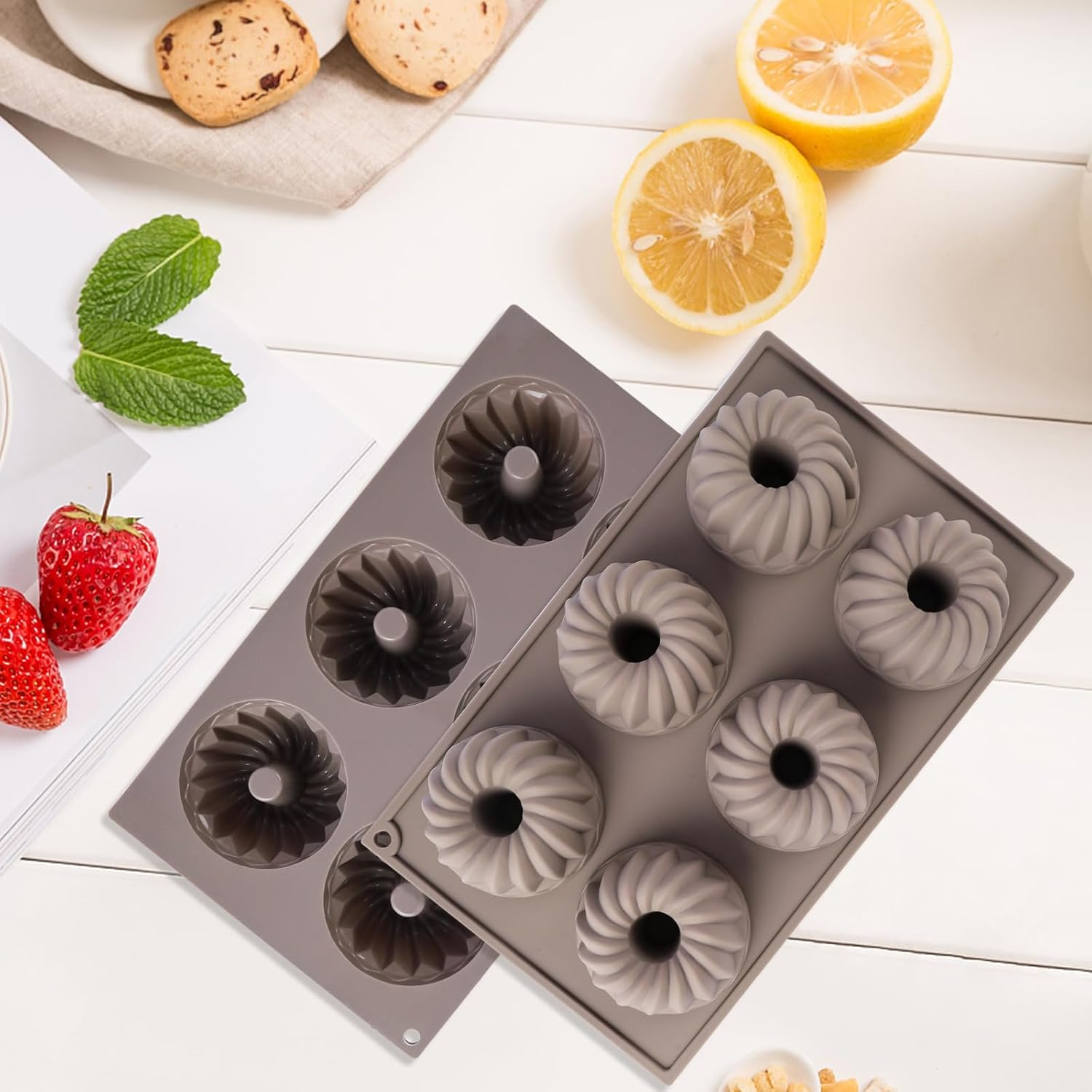 Graue Silikon-Backformen auf weißem Tisch, umgeben von Zitronen, Erdbeeren, Minze und Keksen.