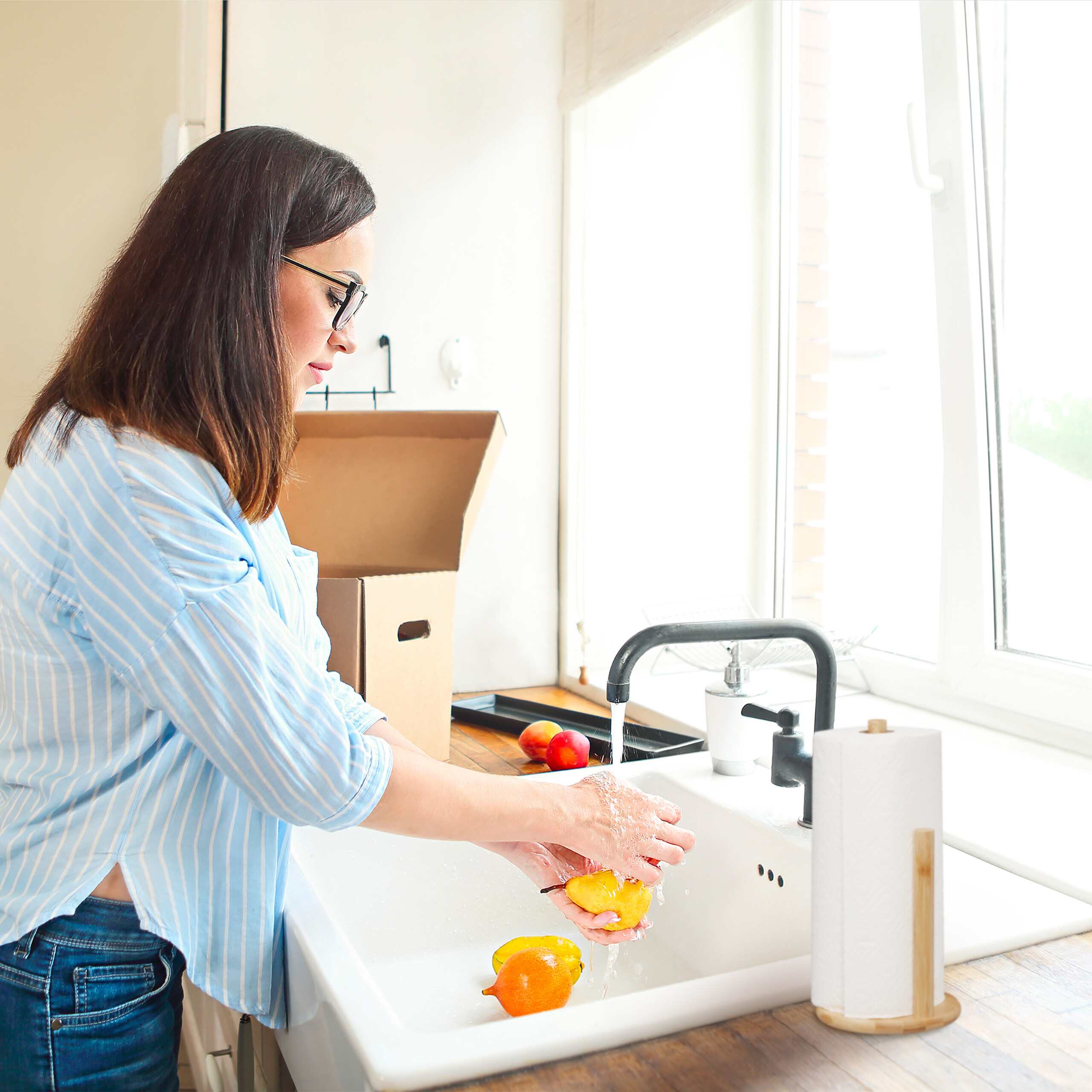Eine Frau mit Brille wäscht gelbe und orange Früchte in einer weißen Spüle am Fenster.