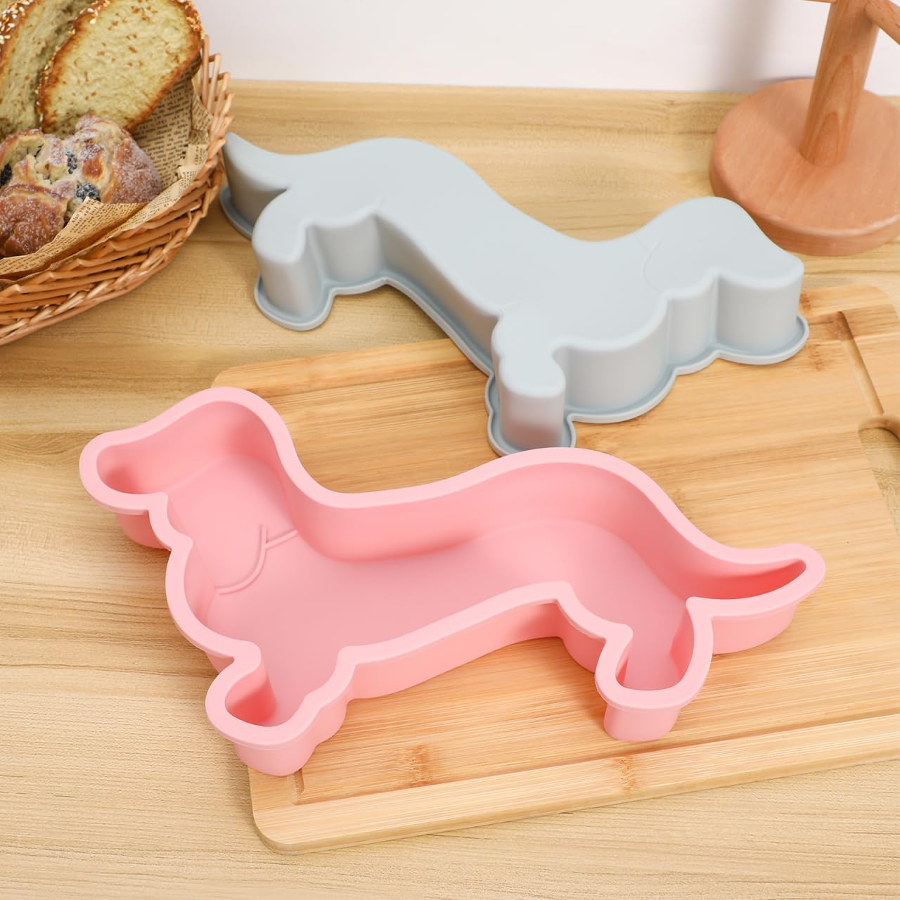 Zwei Dackel-förmige Silikonbackformen, eine rosa und eine graue, auf einem Holzschneidebrett mit Brot daneben.