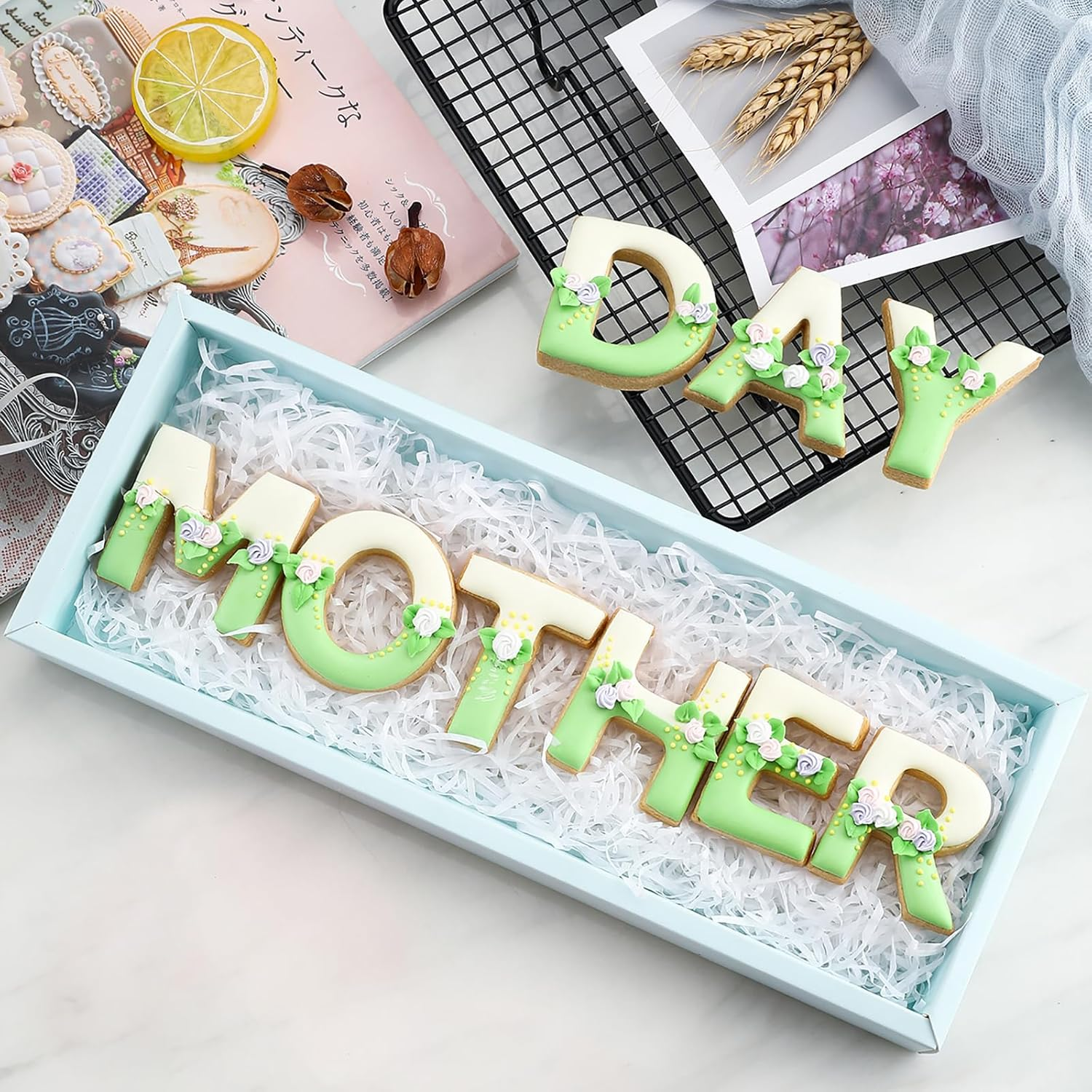 Geschenkkiste mit grün-weißen Plätzchen, die das Wort MOTHER bilden, auf weißem Hintergrund mit Zeitschriften.