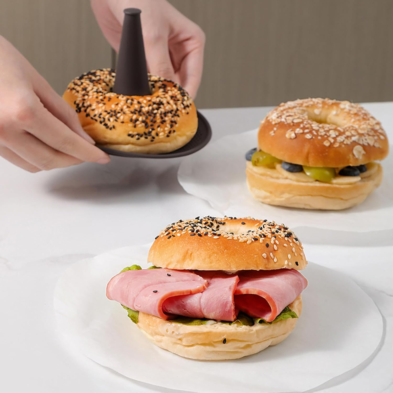 Person nimmt einen Sesam-Bagel von einem Halter ab, zwei weitere Sandwiches mit Schinken und Obst liegen auf dem Tisch.