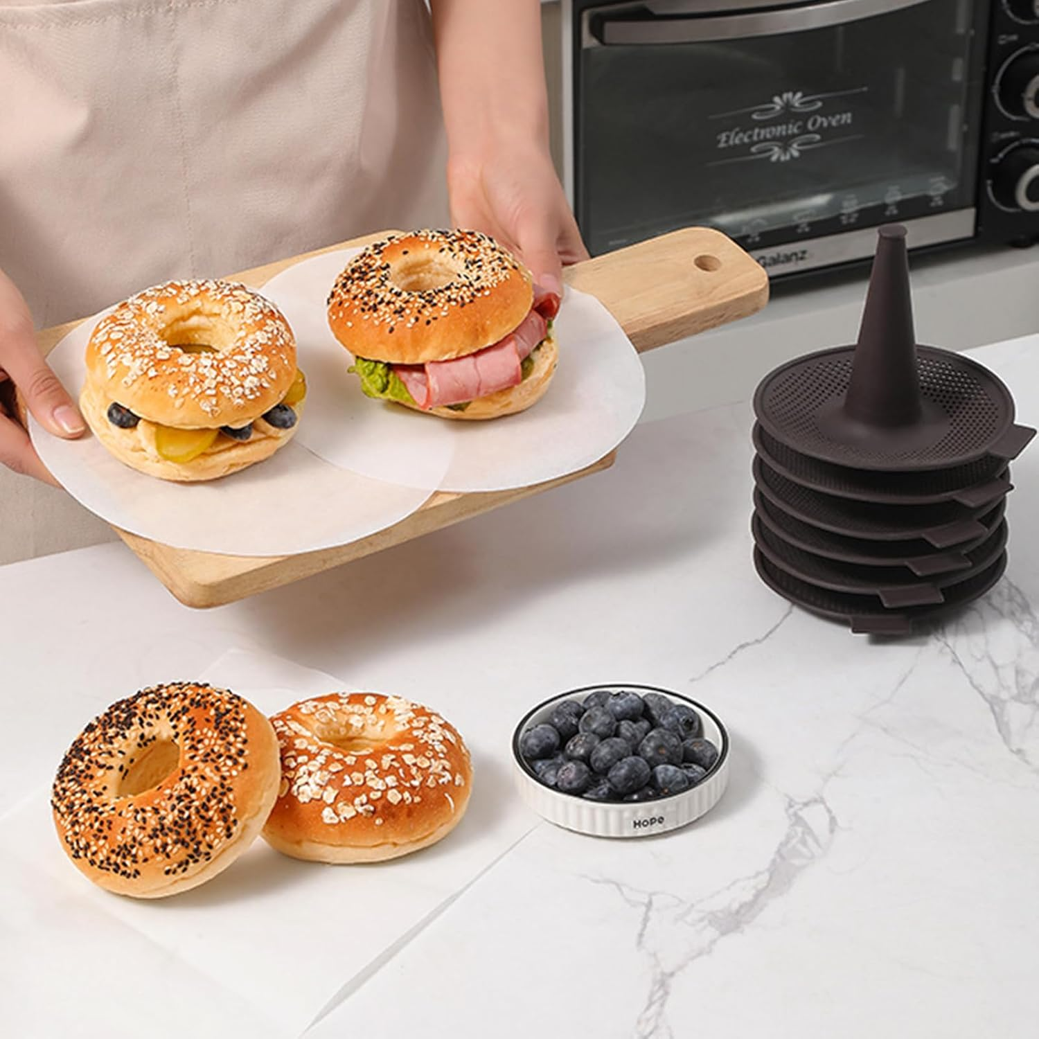Eine Person hält ein Holzbrett mit belegten Bagels, daneben liegen frische Bagels und Blaubeeren.
