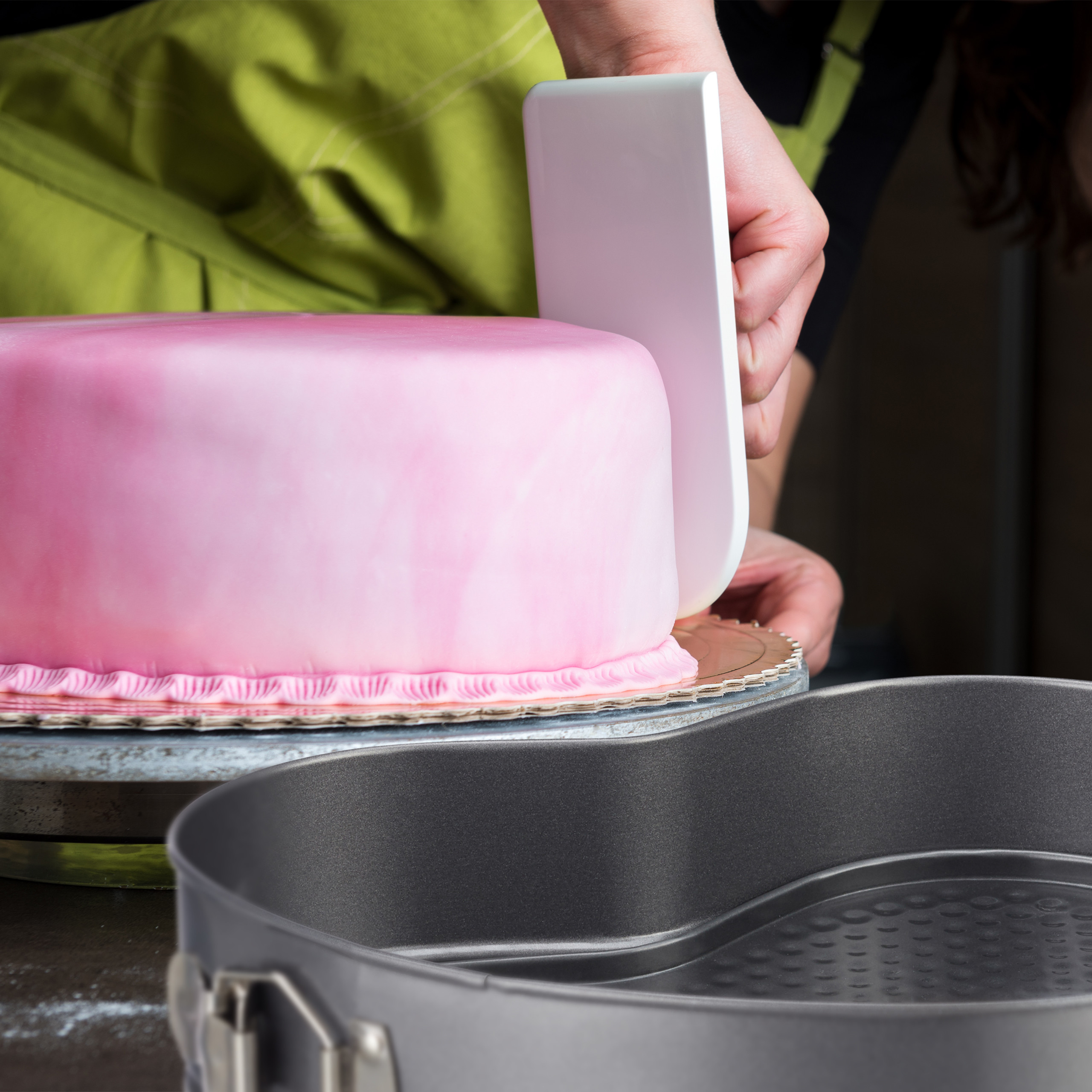 Eine Person glättet rosa Fondant mit einem weißen Schaber auf einem runden Kuchen, im Vordergrund eine Herzform.