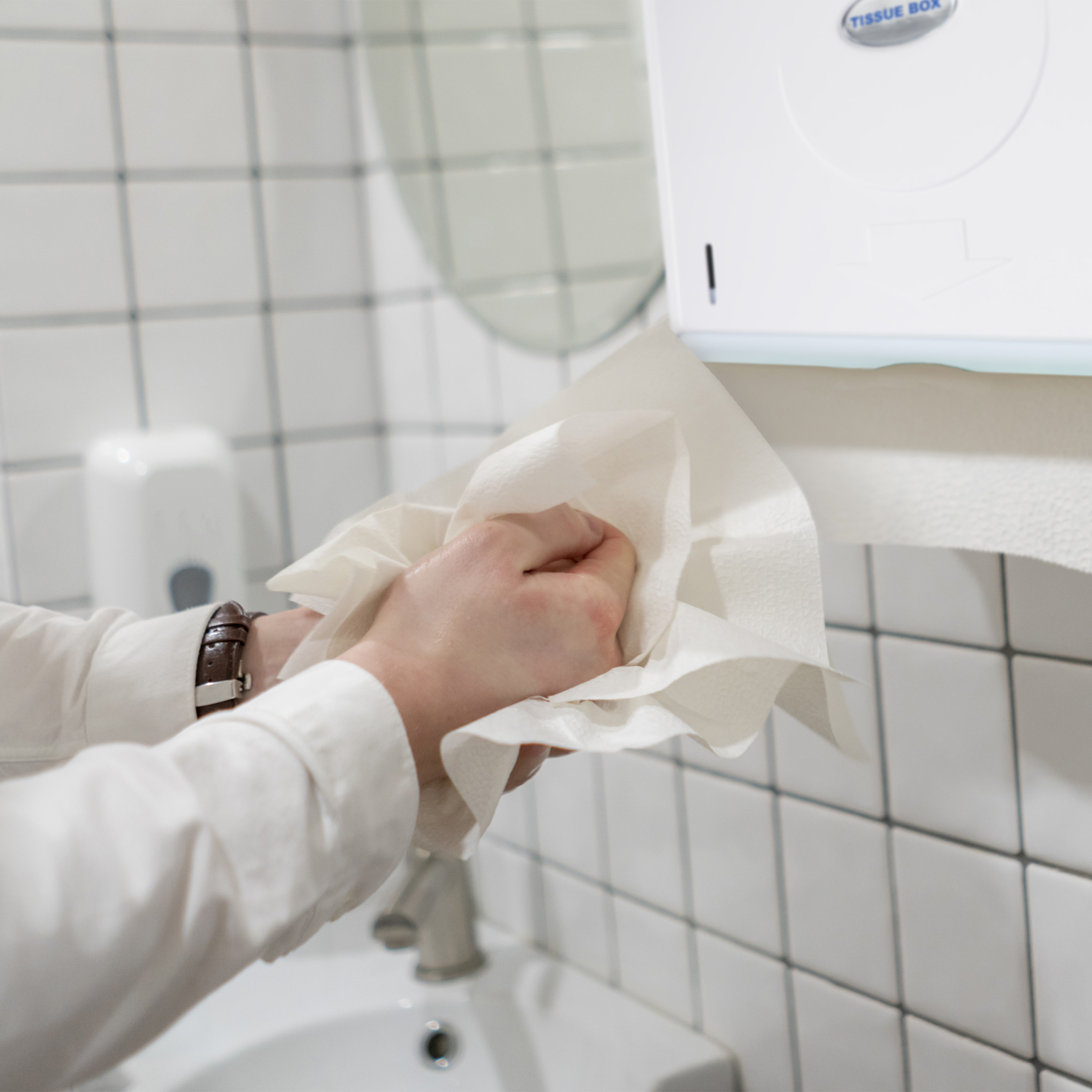 Person trocknet Hände mit Papierhandtuch vor weißem Fliesenhintergrund.