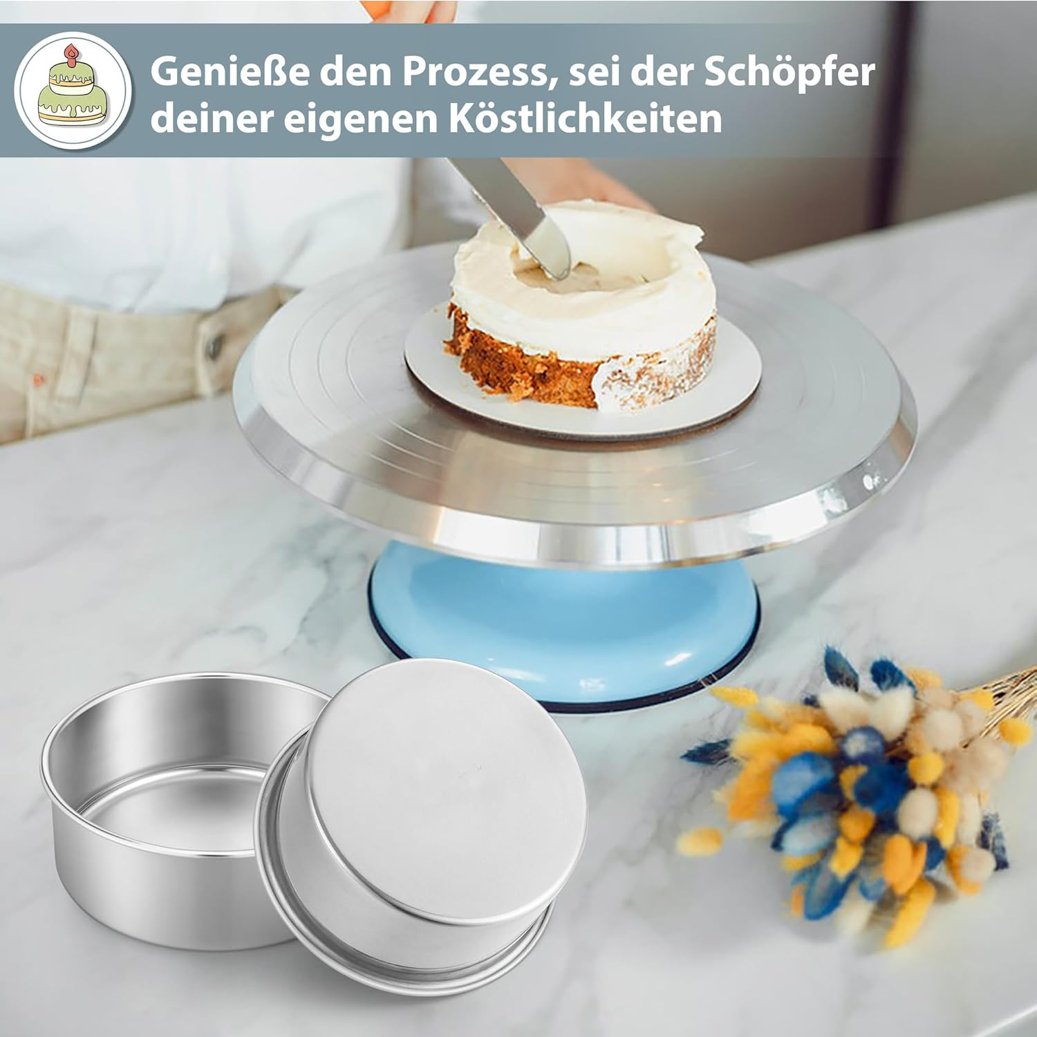 Eine Person bestreicht einen kleinen Kuchen auf einer blauen Drehscheibe. Zwei Metallbackformen stehen auf dem Tisch.