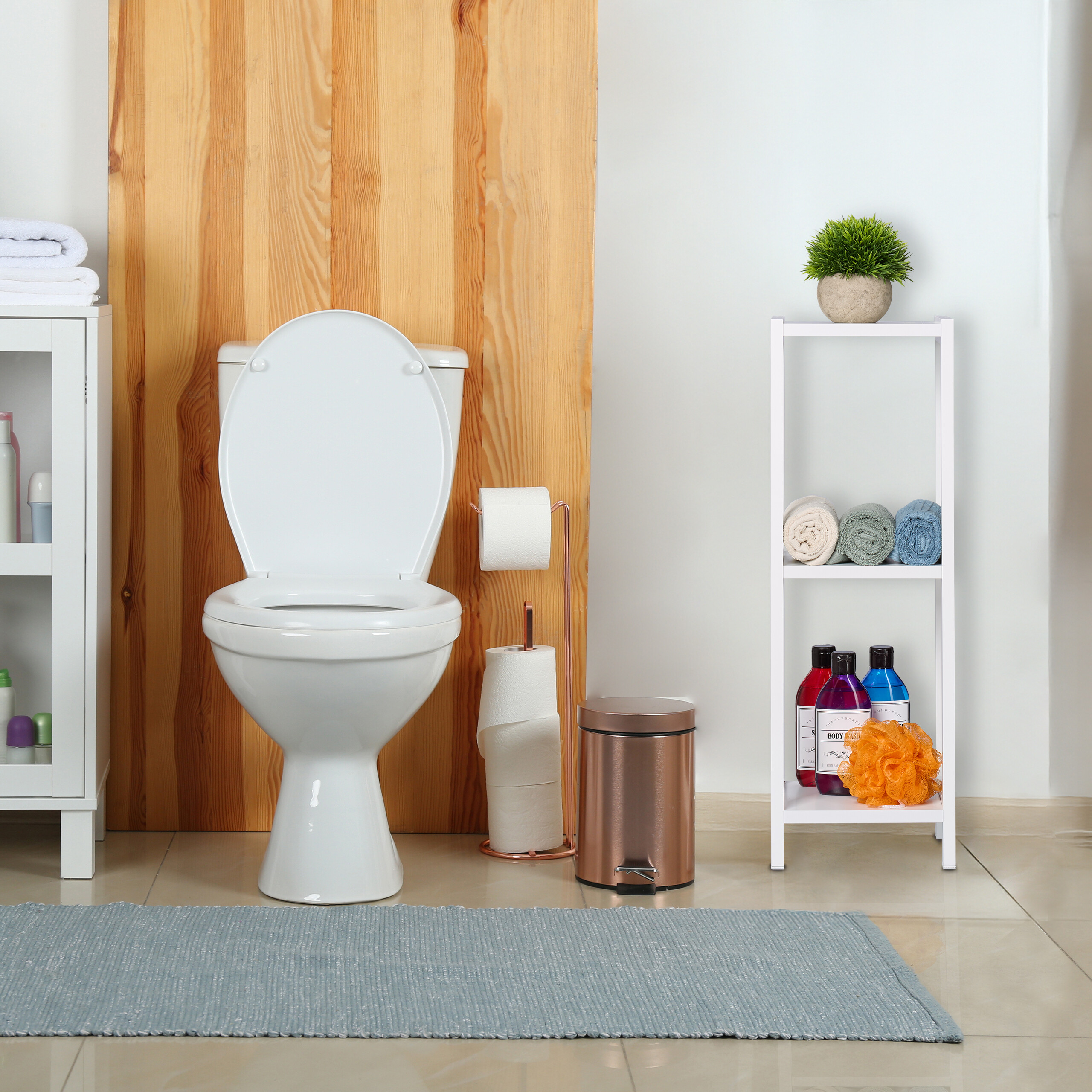 Eine weiße Toilette steht vor einer holzverkleideten Wand, daneben ein Regal mit Badutensilien und ein Mülleimer.