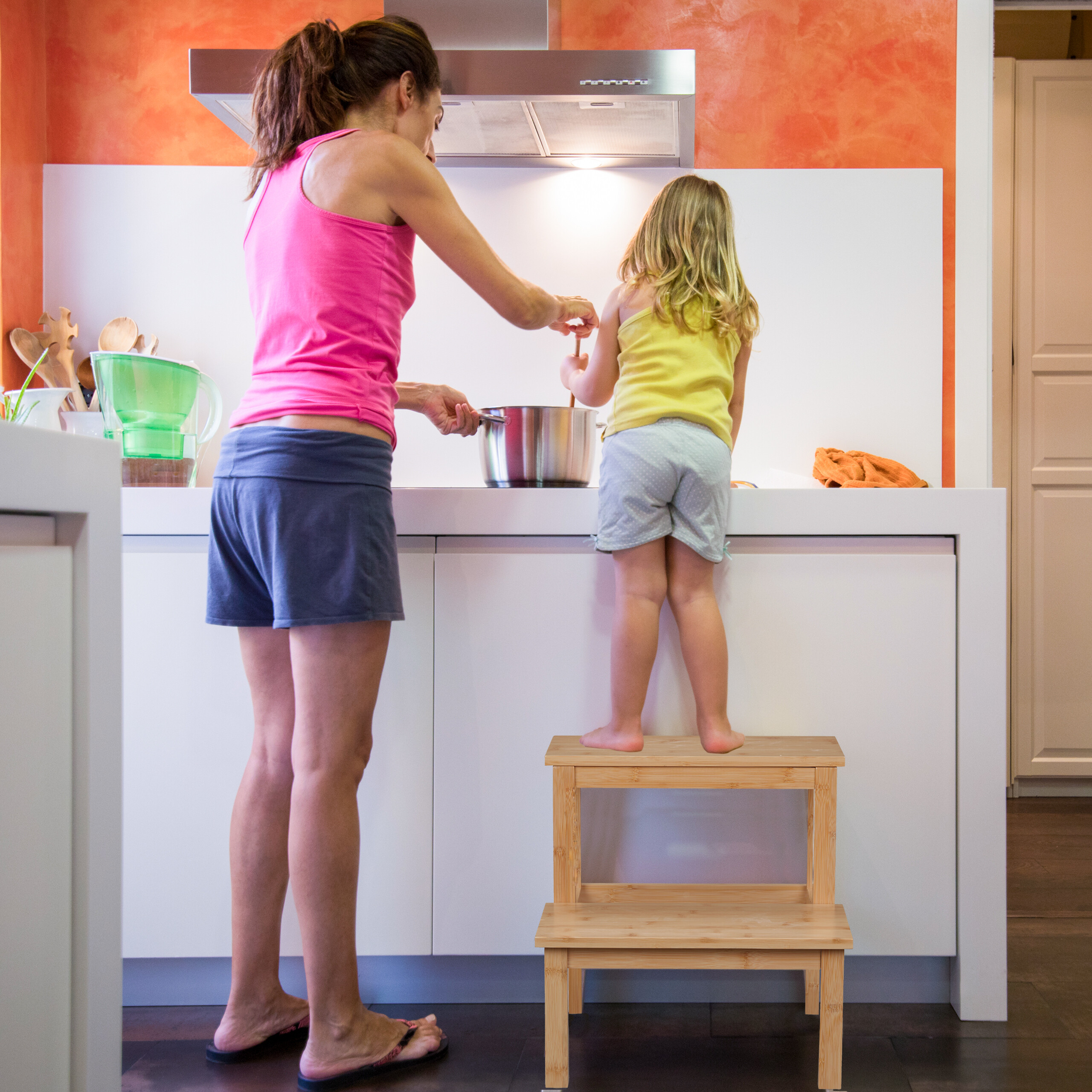 Eine Frau in Pink und ein Mädchen auf einem Hocker kochen gemeinsam am Herd in einer modernen Küche.