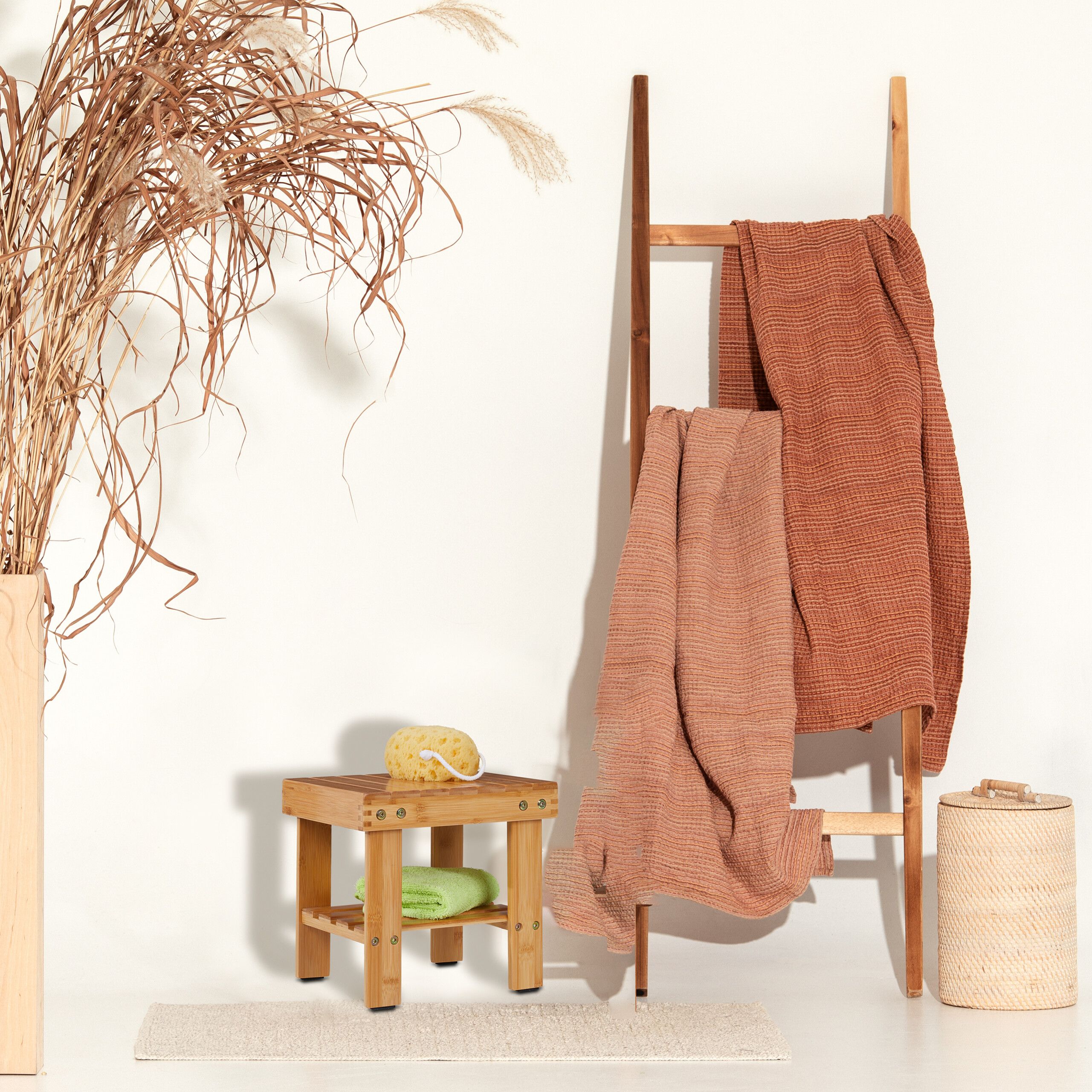 Eine Holzlade hält braune Handtücher neben einem Hocker, einem Korb und Gras in einer Vase vor weißer Wand.