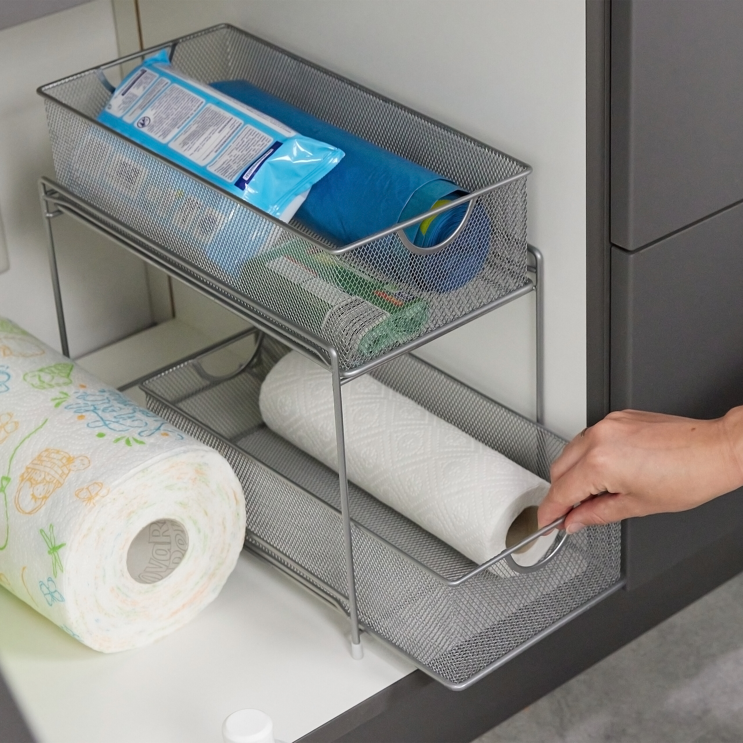 Eine Hand zieht einen zweistöckigen Drahtkorb-Organizer, gefüllt mit Küchenpapier und Reinigungsmitteln.