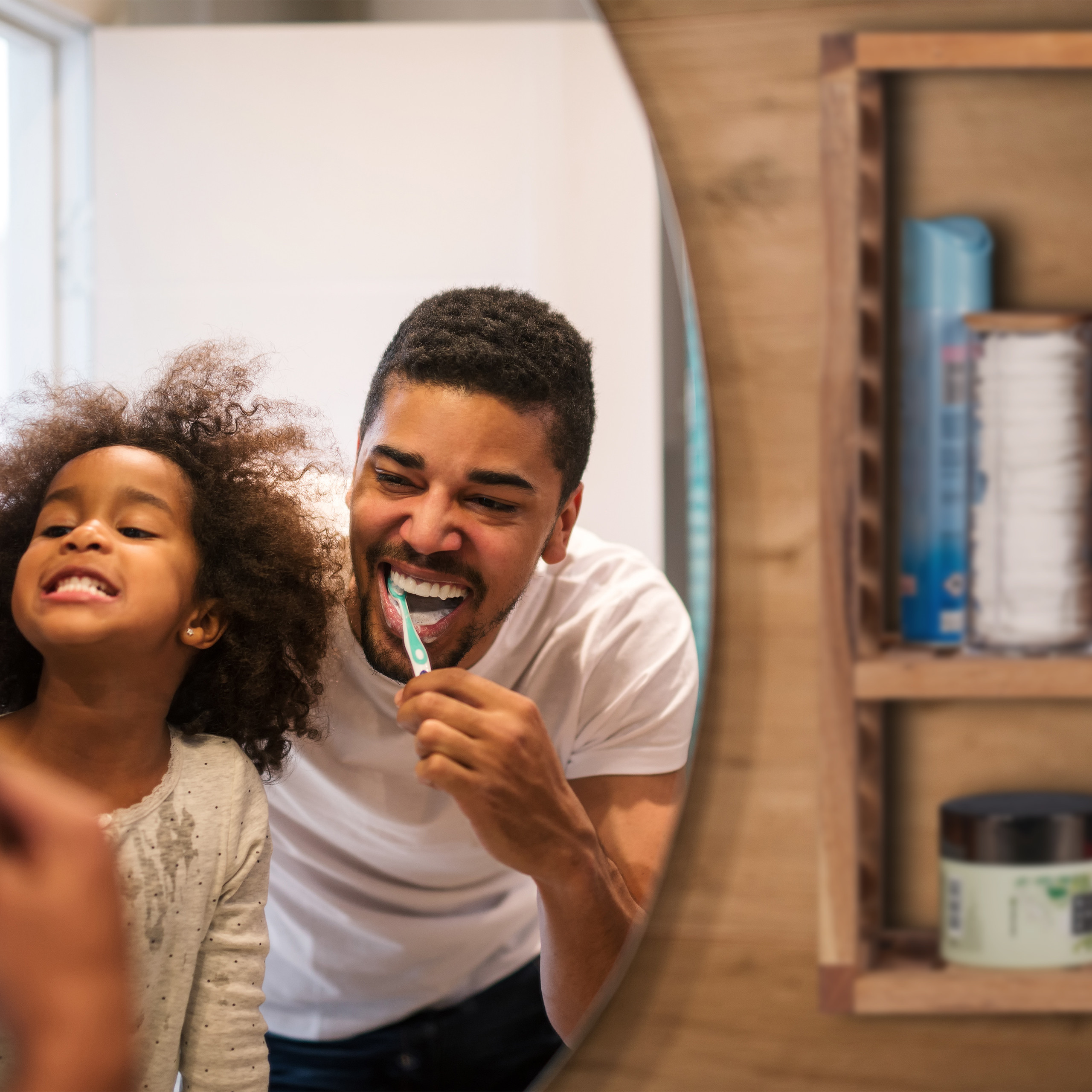 Ein Vater und seine kleine Tochter putzen gemeinsam lächelnd ihre Zähne vor einem Badezimmerspiegel.