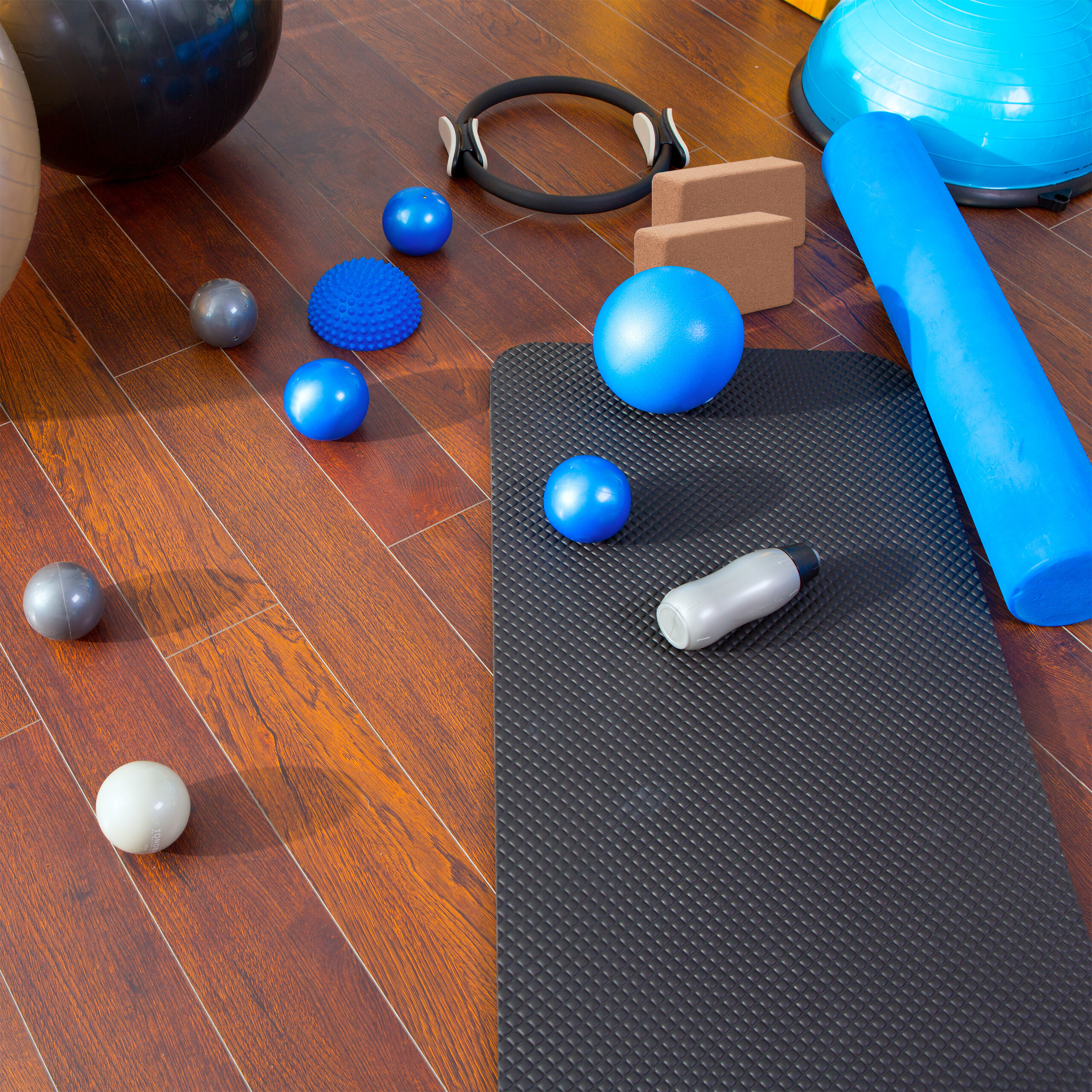 Verschiedene Pilates- und Fitnessgeräte wie Matten, Bälle, Schaumstoffrollen und Blöcke liegen auf Holzboden.