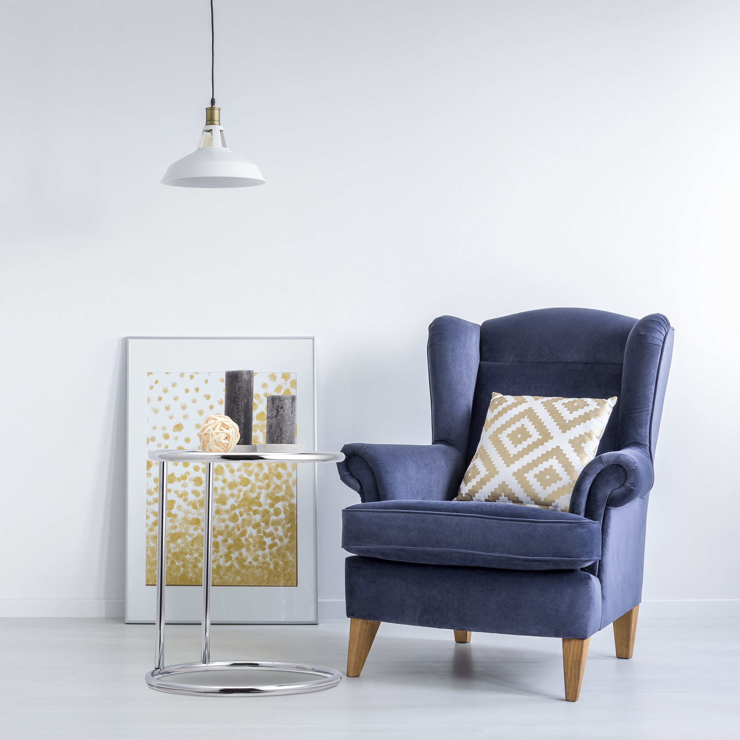 Ein blauer Sessel mit Kissen, ein Beistelltisch, gerahmte Kunst und eine Hängelampe vor weißer Wand.