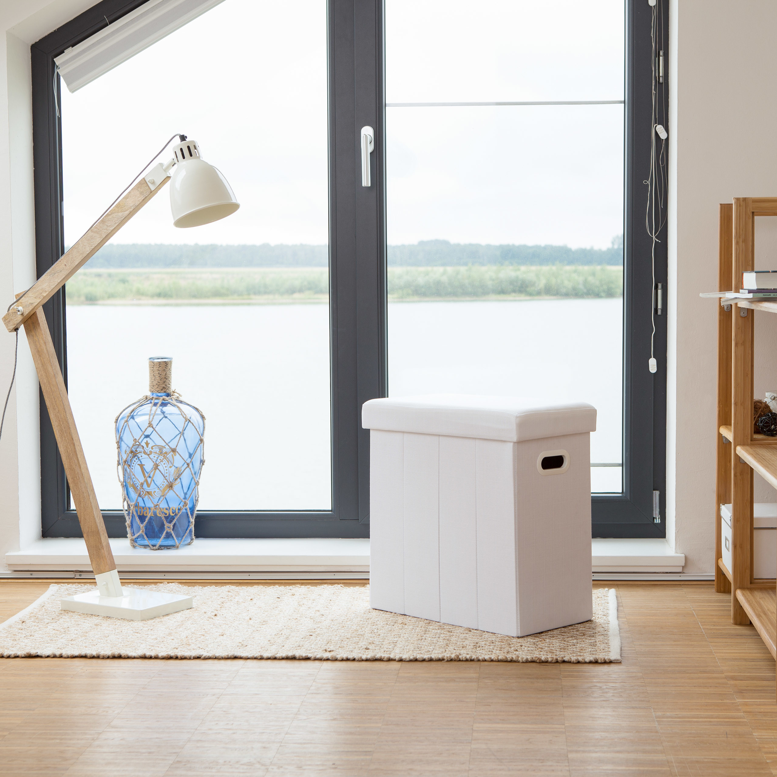 Eine weiße Aufbewahrungsbox, Holzstehlampe und blaue Glasflasche stehen vor einem großen Fenster mit Seeblick.