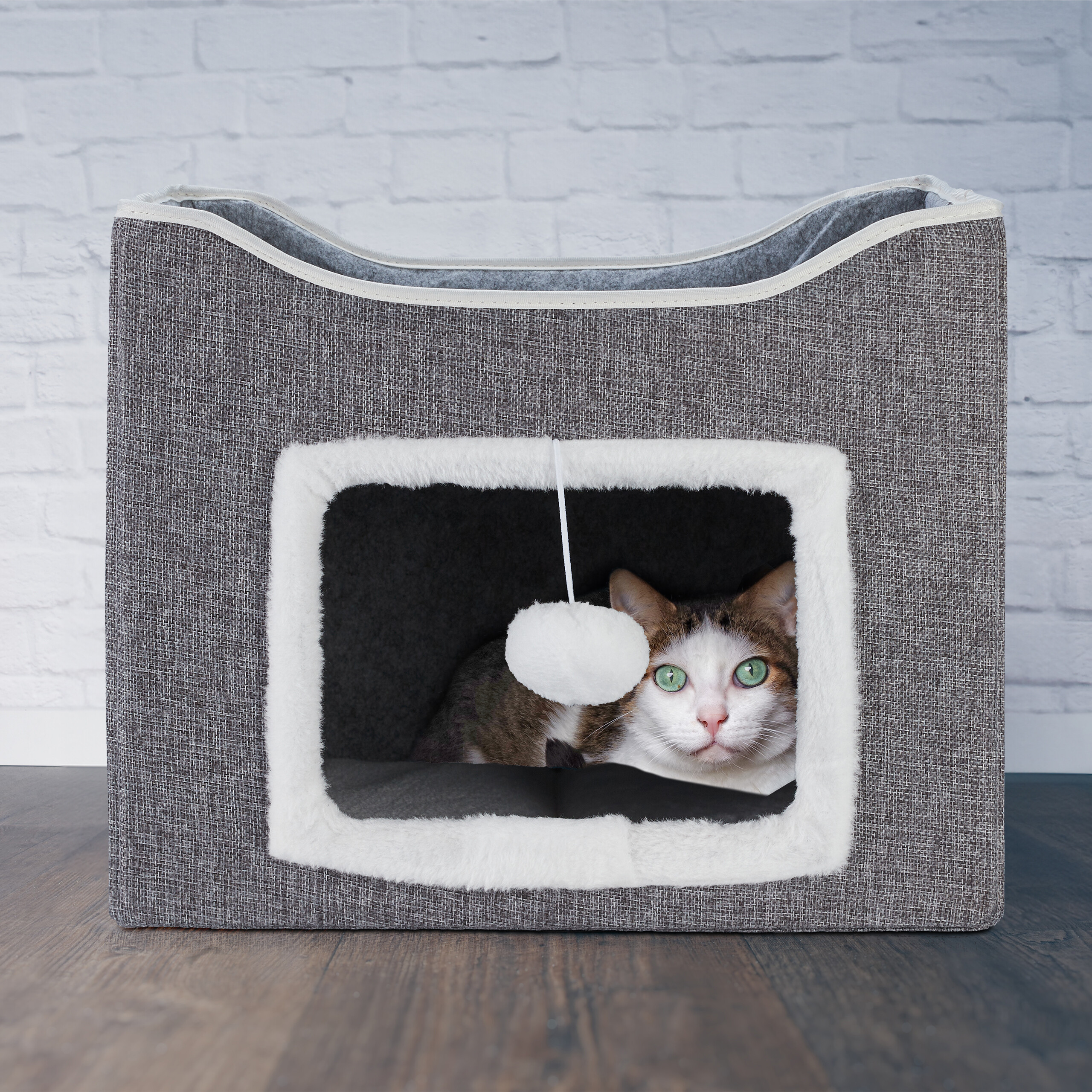 Eine Katze mit grünen Augen schaut aus einem grauen Stoff-Katzenhaus mit weißem Plüscheingang.