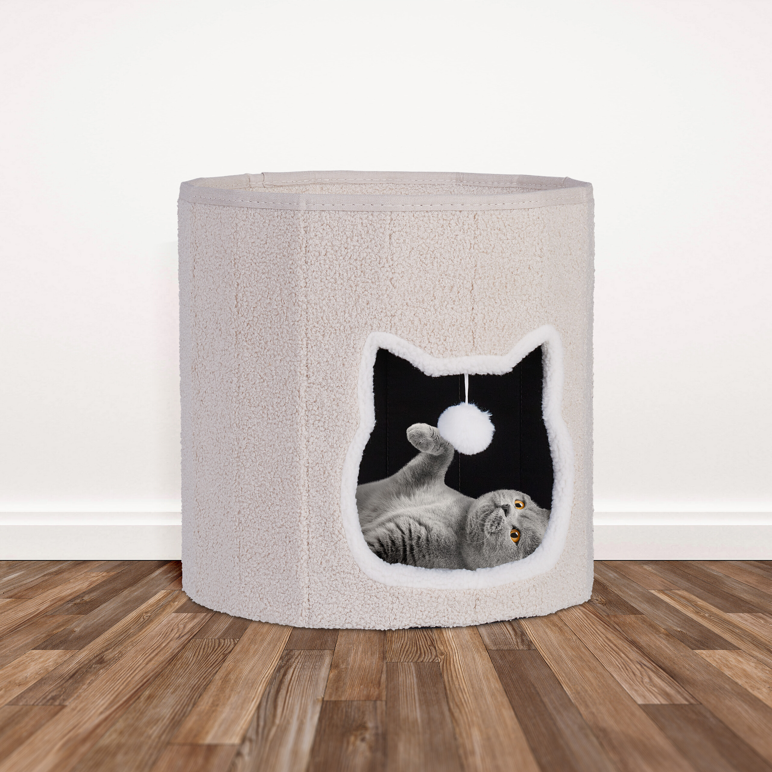Eine graue Katze spielt mit einem baumelnden Pompon in einem beigen Katzenhaus mit Katzen-Tür auf Holzfussboden.