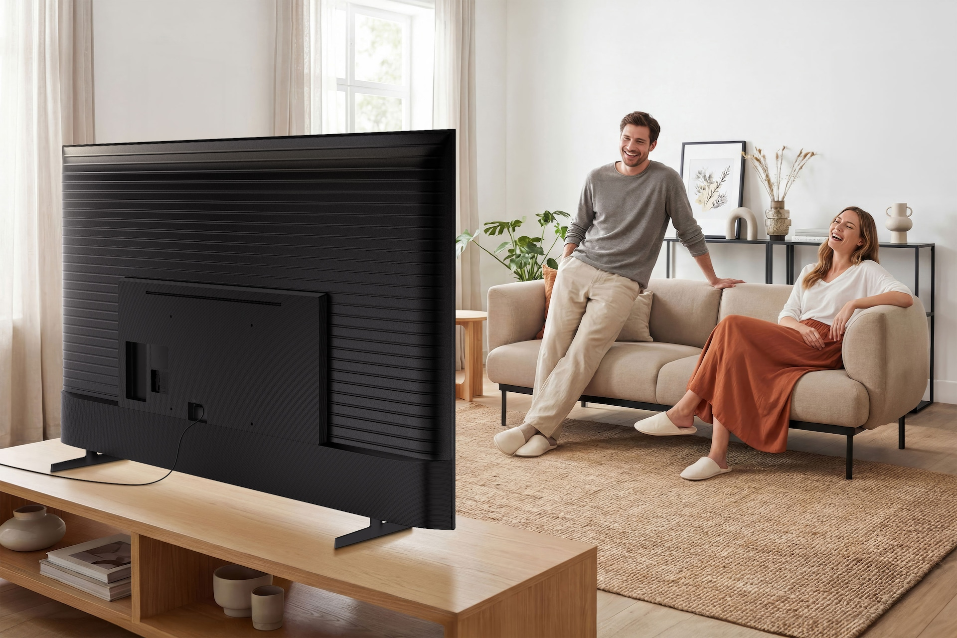 Un gran televisor negro sobre una mesa de madera, mientras una pareja al fondo está sentada en un sofá.